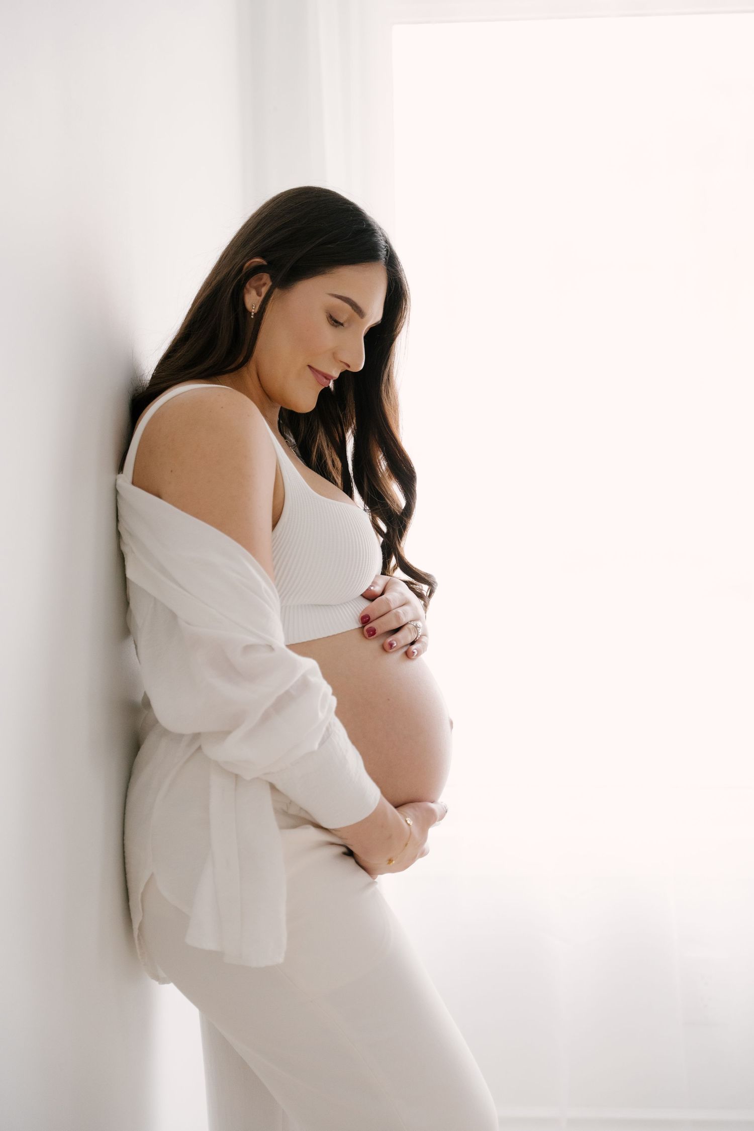 Serene pregnant woman in white open shirt and bra cradling her bare baby bump against a bright wall.