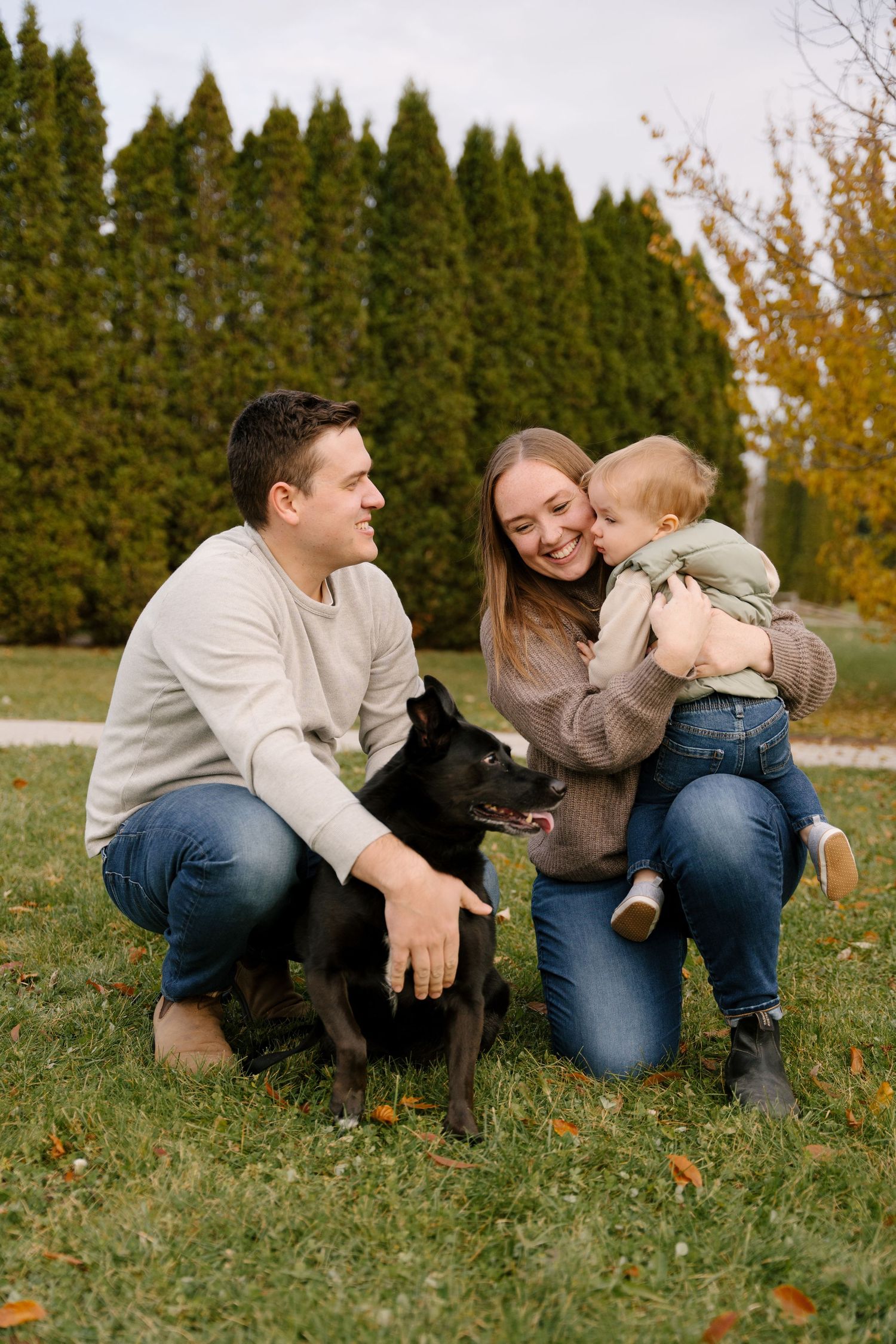 Smiling family with baby and black dog posing together on grass in fall park.