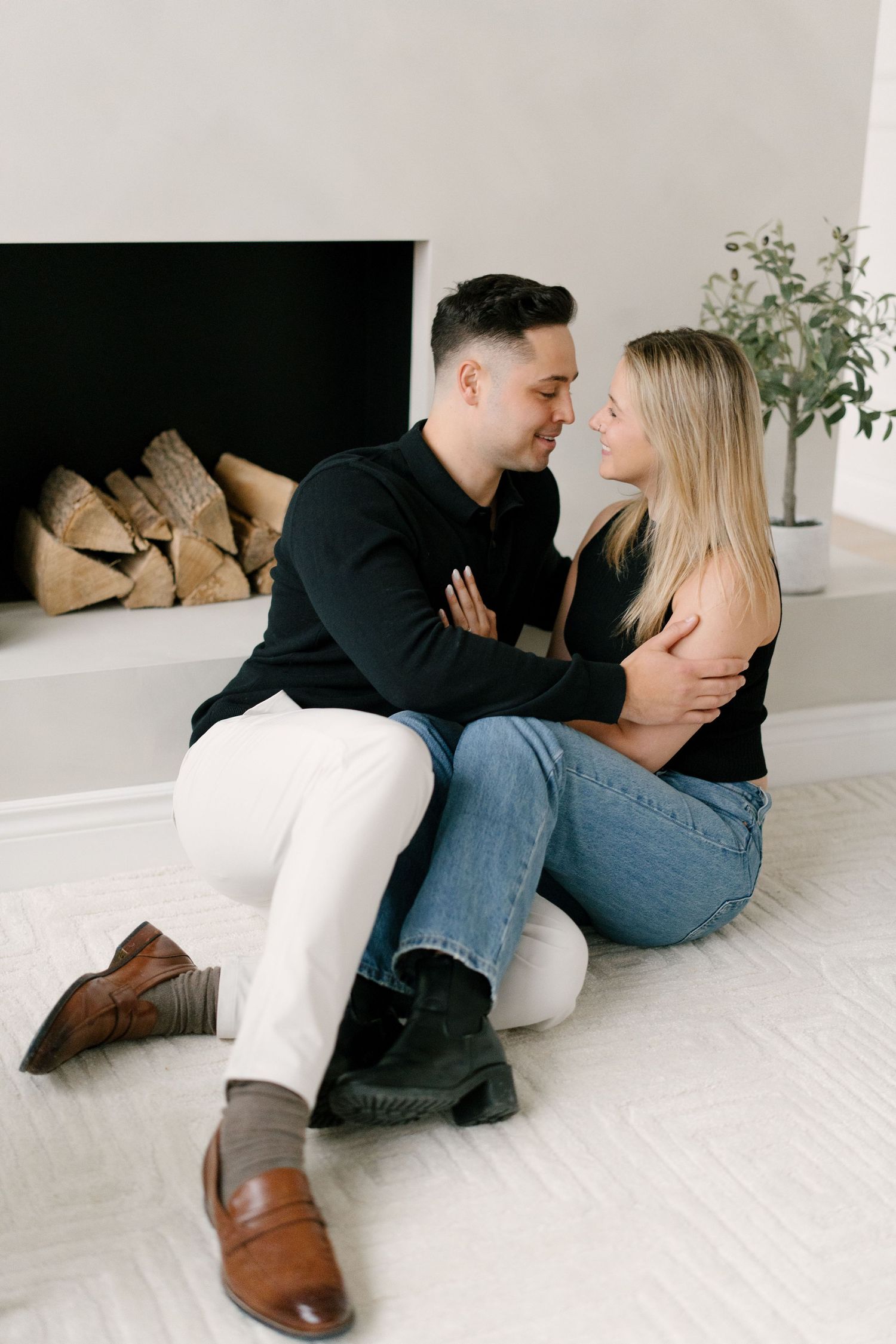 Couple sitting close together near a modern fireplace with stacked logs in a bright white room.