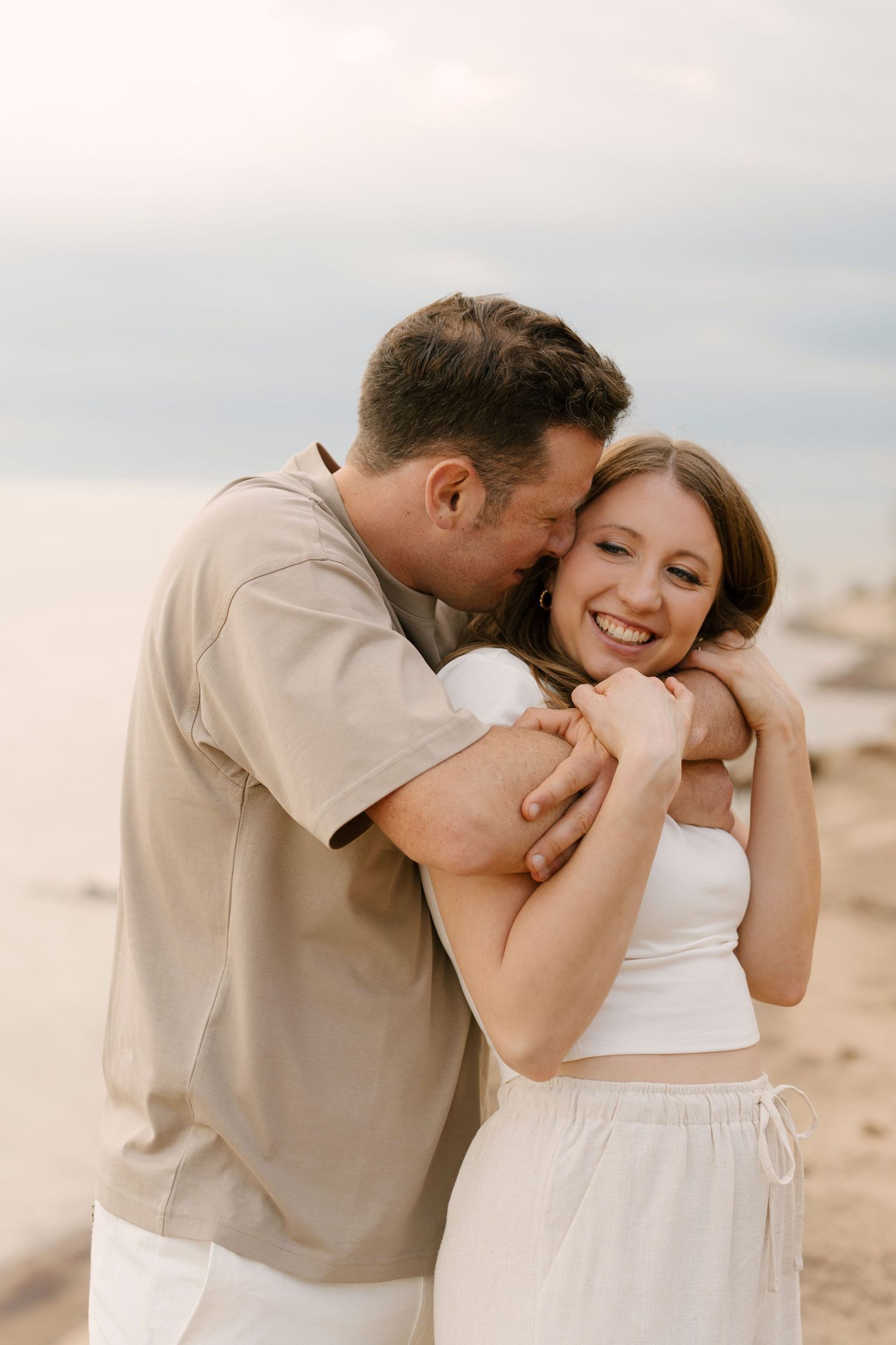 Man hugging woman from behind on beach, both smiling joyfully near the water.