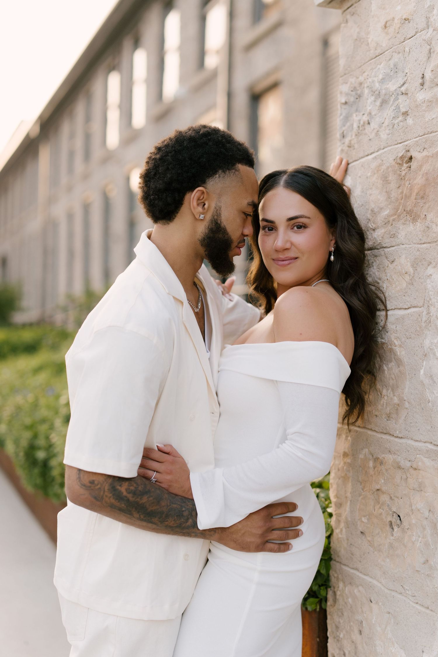 Couple in white outfits embrace against a stone wall, woman in off-shoulder dress smiles at camera.