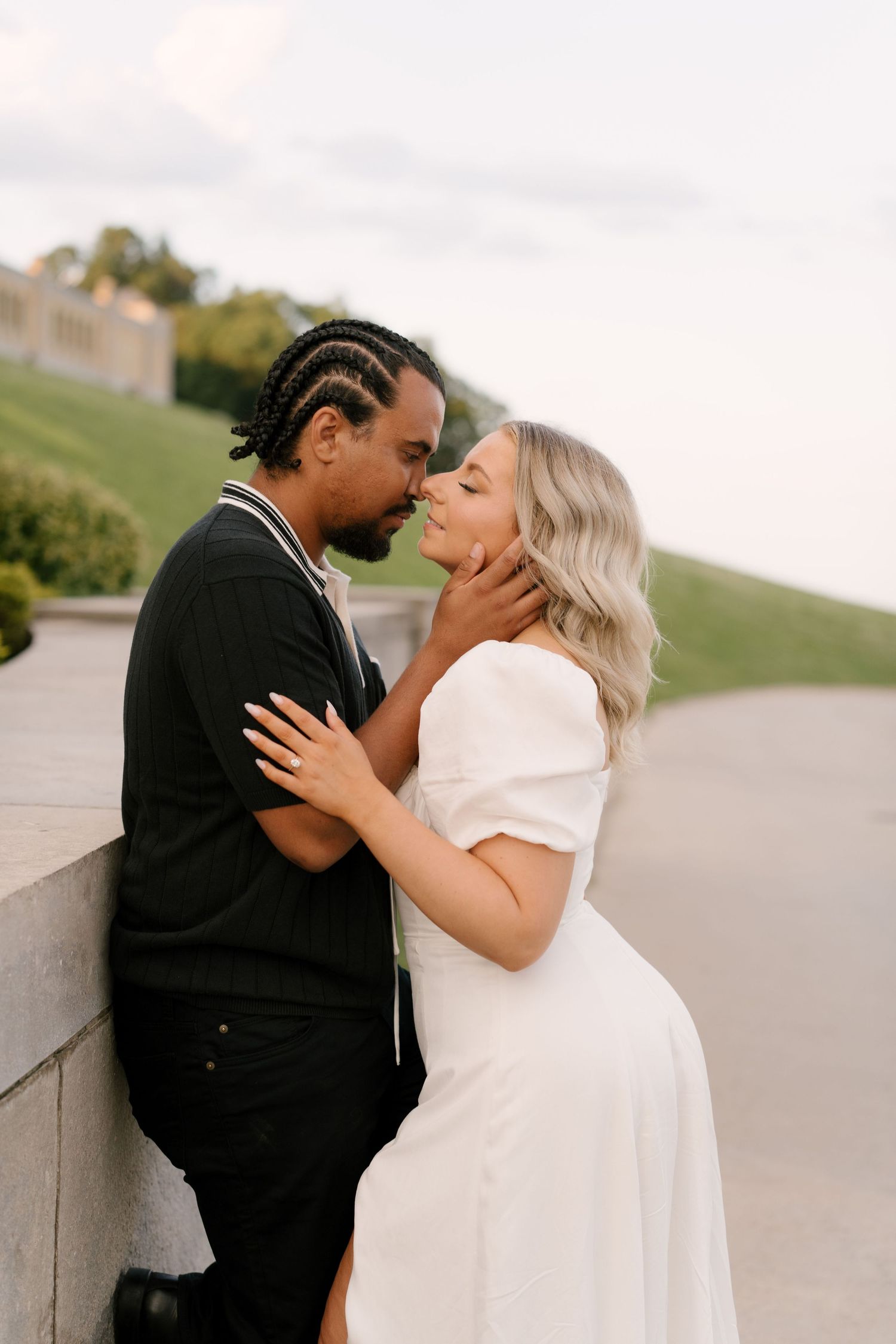 Couple sharing an intimate almost-kiss moment leaning against a stone wall at dusk outdoors.
