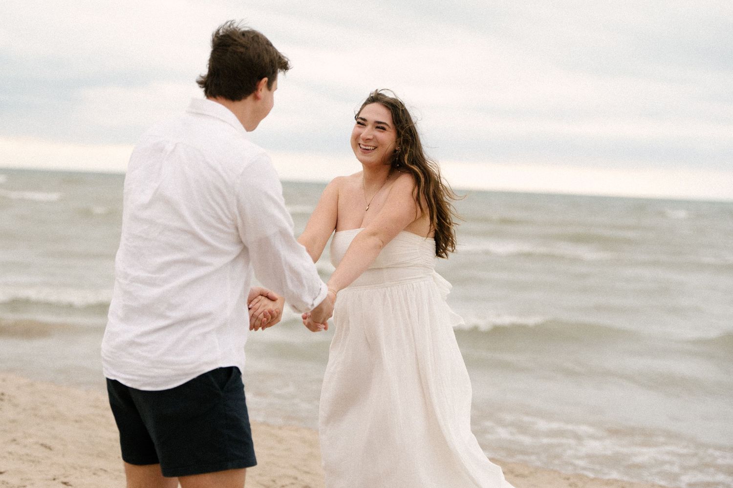 Smiling bride in white strapless dress holding hands with groom on a windy beach shoreline.
