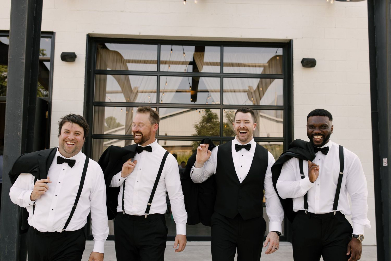 Four groomsmen in black tuxedos with suspenders laughing together outside a St Marys wedding venue.