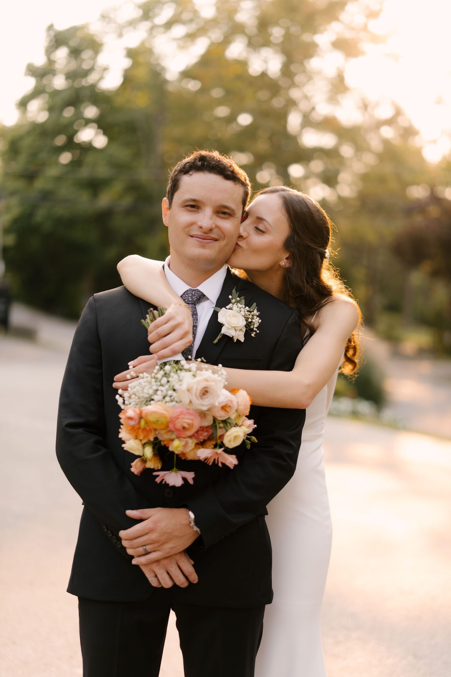 Bride kisses groom on cheek while holding colorful bouquet outdoors at golden hour wedding portrait.