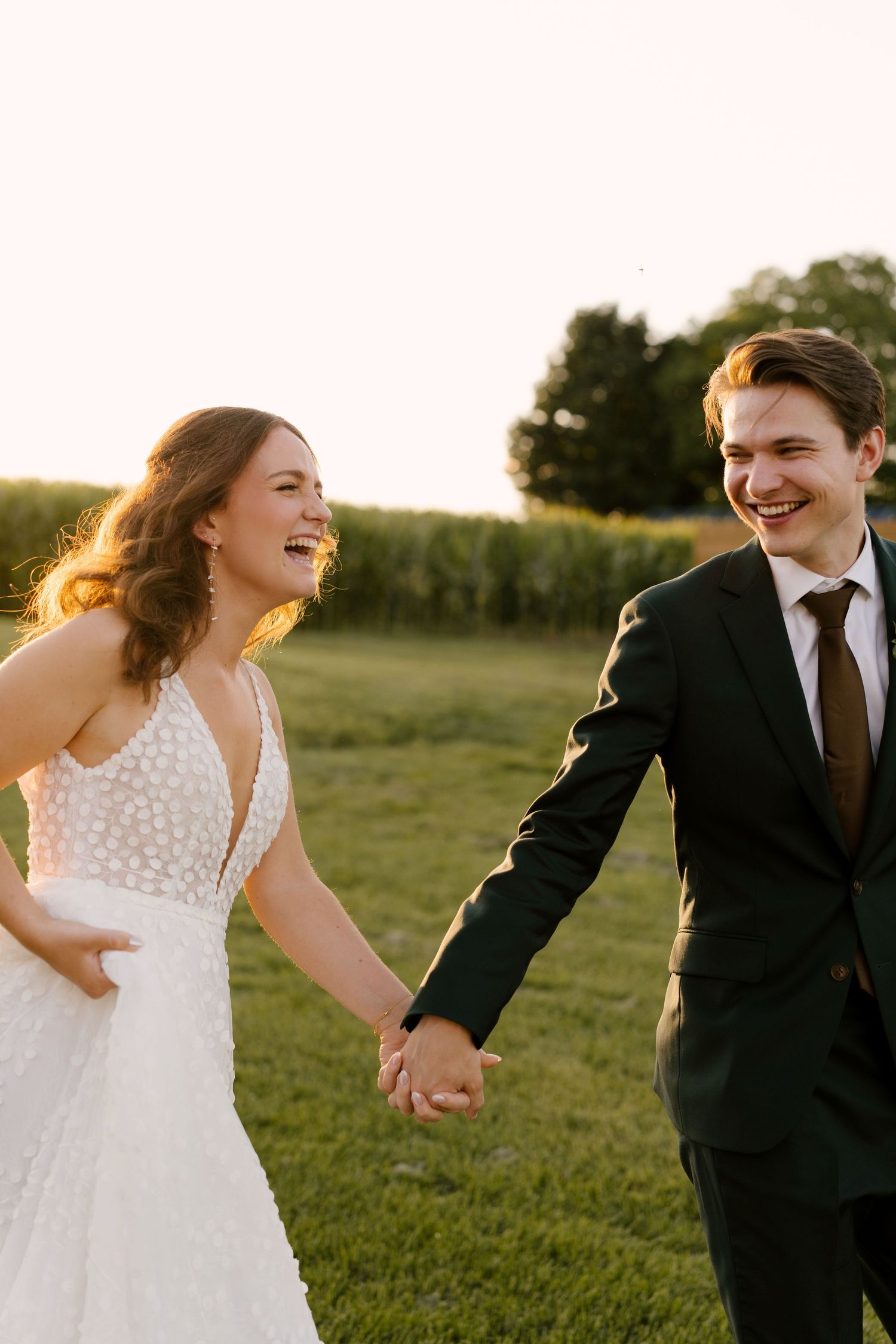 Happy bride and groom holding hands and laughing while walking through a sunlit green field.