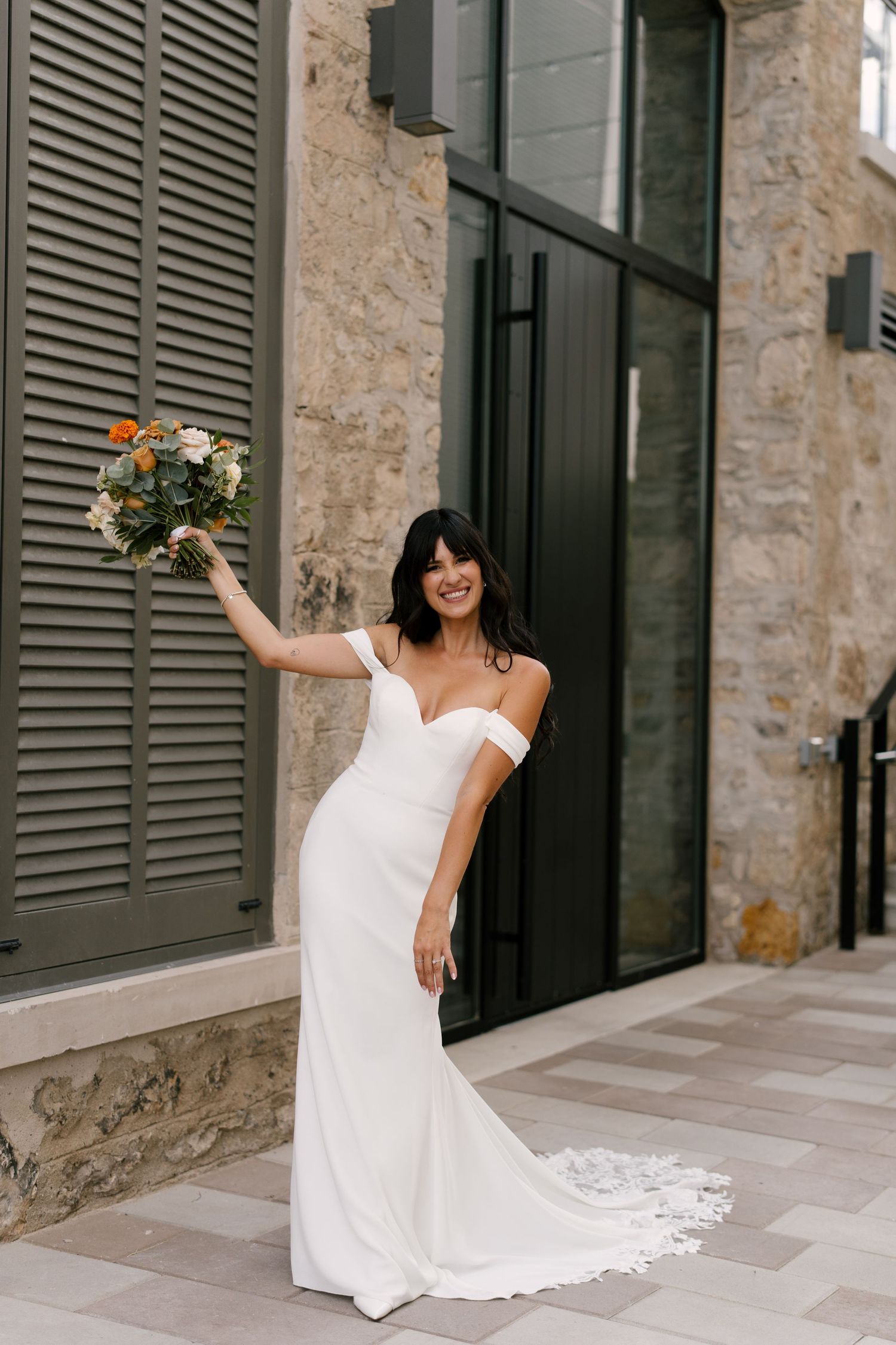 Radiant bride in sleek off-shoulder white gown with lace train raises bouquet triumphantly outside stone Tapestry Hall venue.