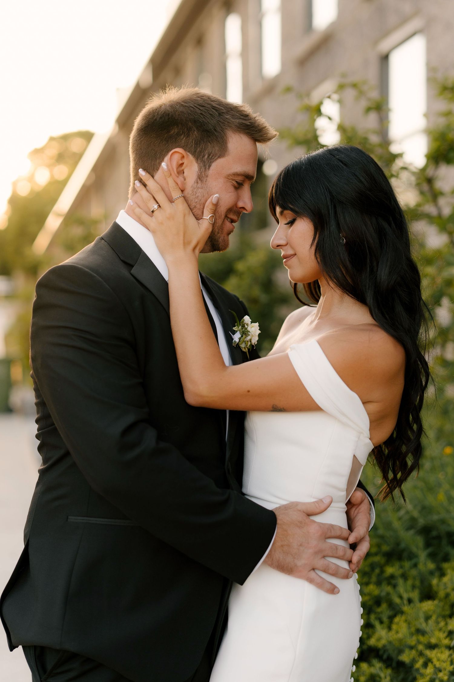 Bride and groom sharing an intimate moment, nearly kissing, bathed in warm golden sunset light outside wedding venue.