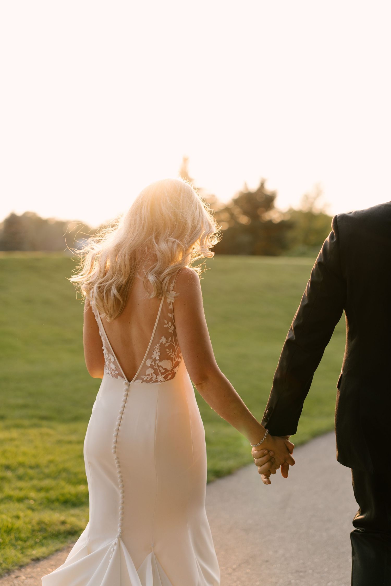 Bride and groom holding hands walking at sunset, bride in backless lace wedding gown with flowing blonde hair.