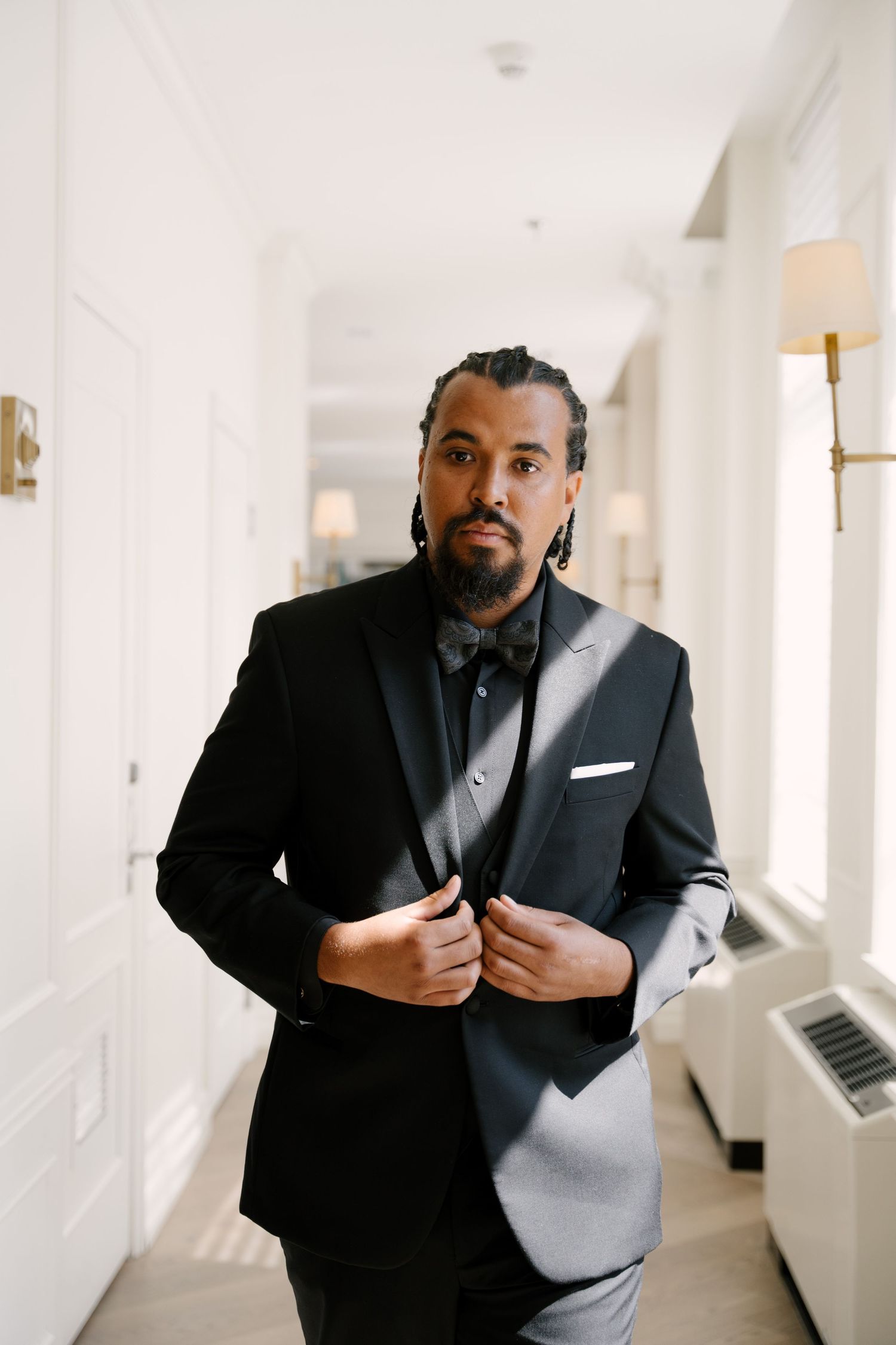Stylish groom in black tuxedo adjusting jacket lapels in a bright hallway at The Walper Hotel.