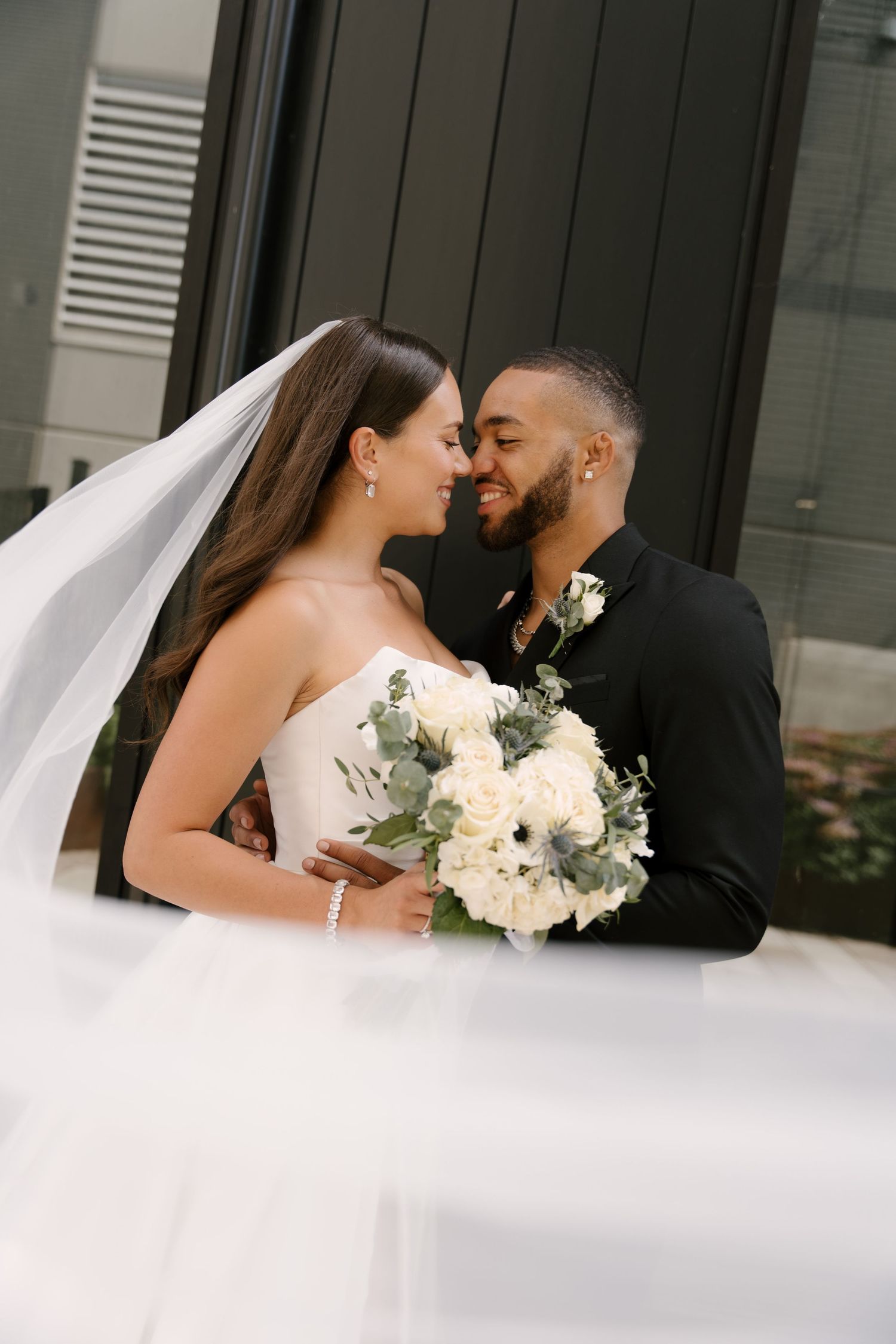 Bride and groom nearly kiss while bride's veil flows in the wind, holding a white floral bouquet outside Tapestry Hall venue.
