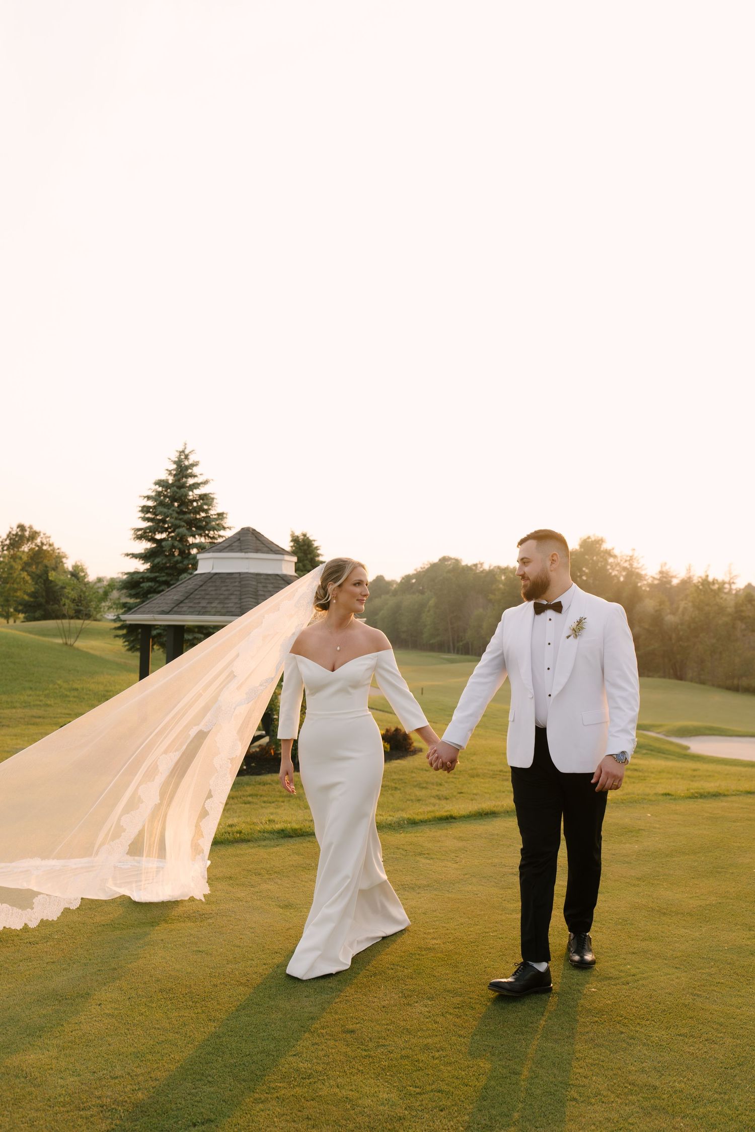 Bride and groom holding hands walking on Whistle Bear golf course at sunset, her lace veil flowing dramatically in the wind.