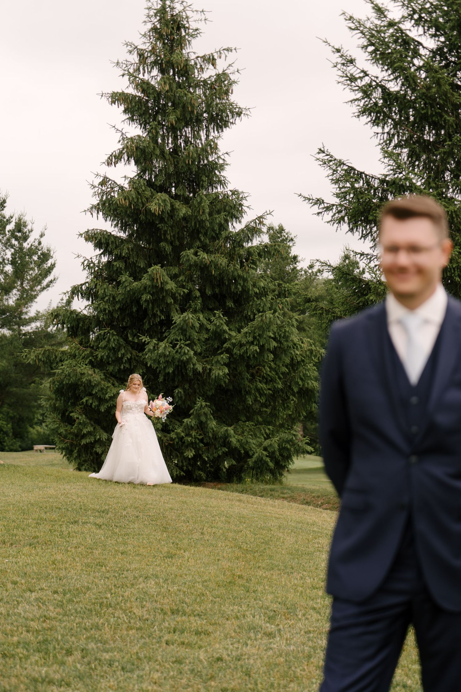 Bride in white gown stands near evergreen trees as groom smiles in the foreground at St Marys Golf Club.