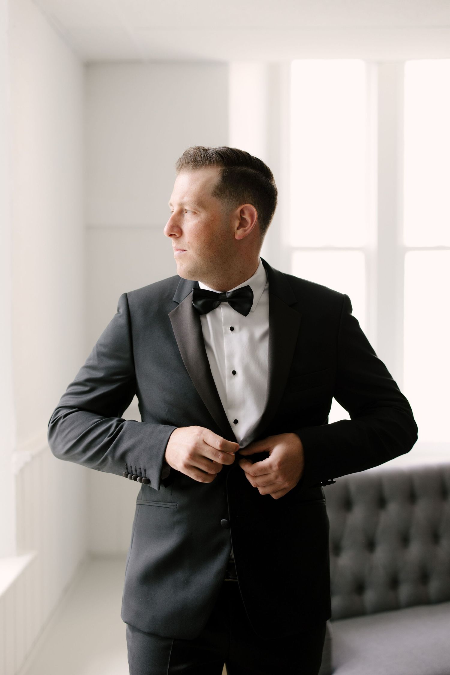 Dapper groom in black tuxedo buttons his jacket while gazing out a bright window before his wedding ceremony.