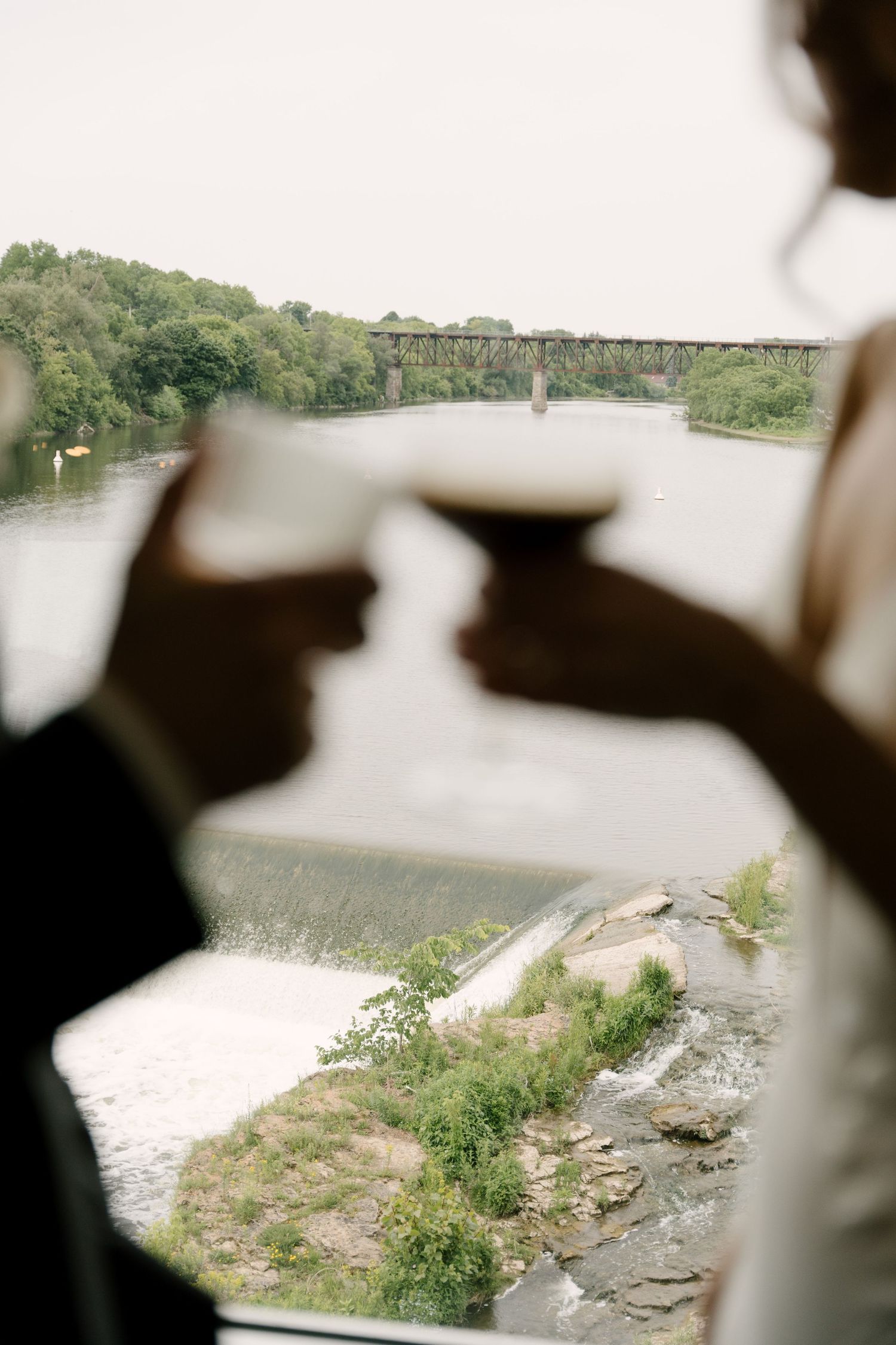 Bride and groom clink glasses in silhouette against panoramic river and bridge backdrop at Cambridge Mill.