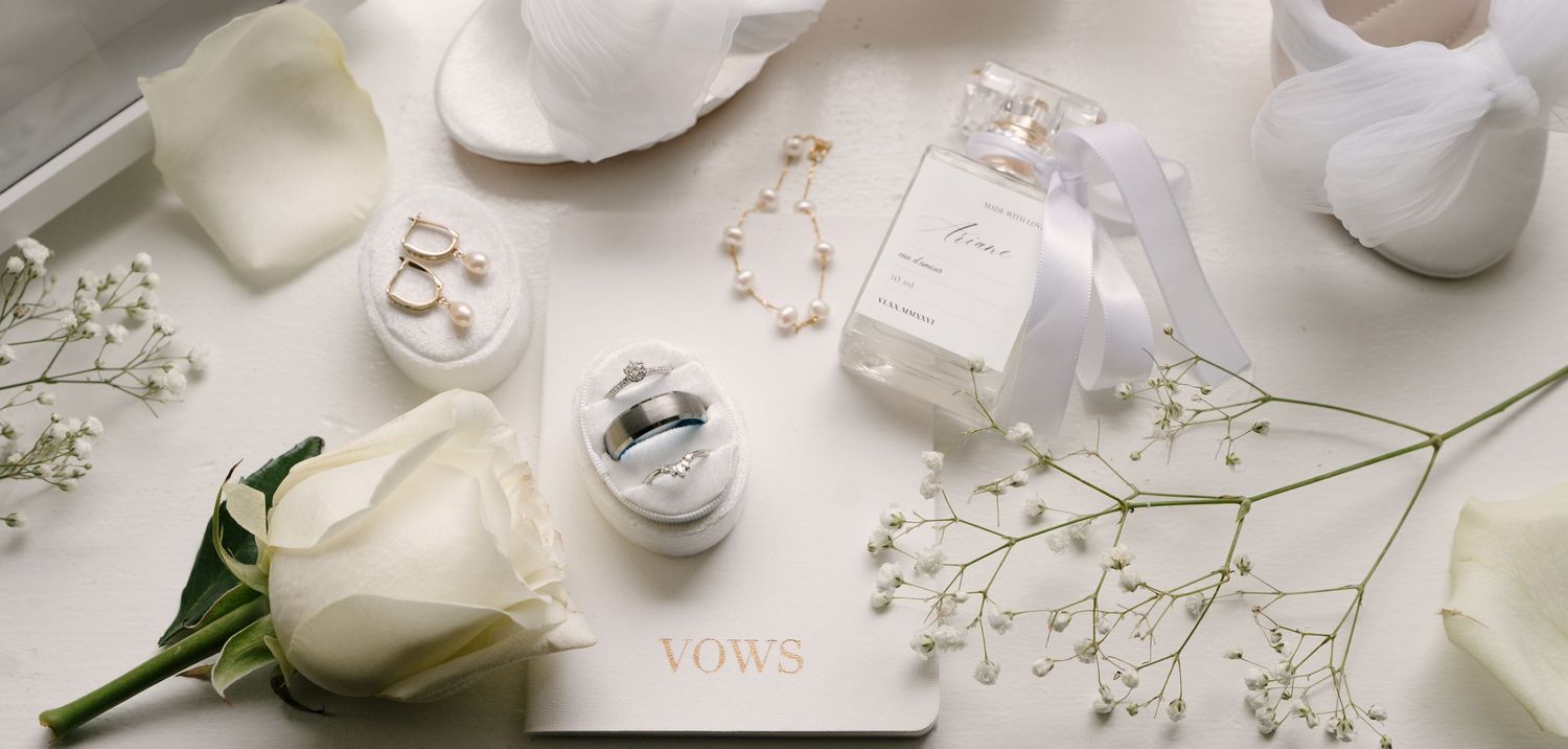 Elegant wedding flat lay with white rose, vow book, rings, pearl bracelet, and gypsophila on white surface.