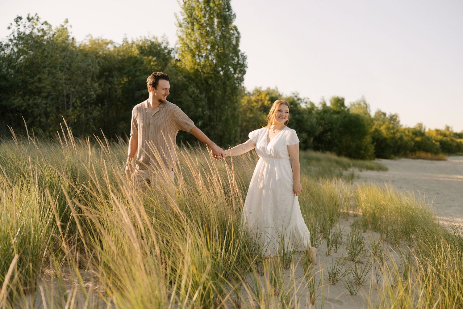 Couple holding hands while walking through sandy beach dunes at golden hour.