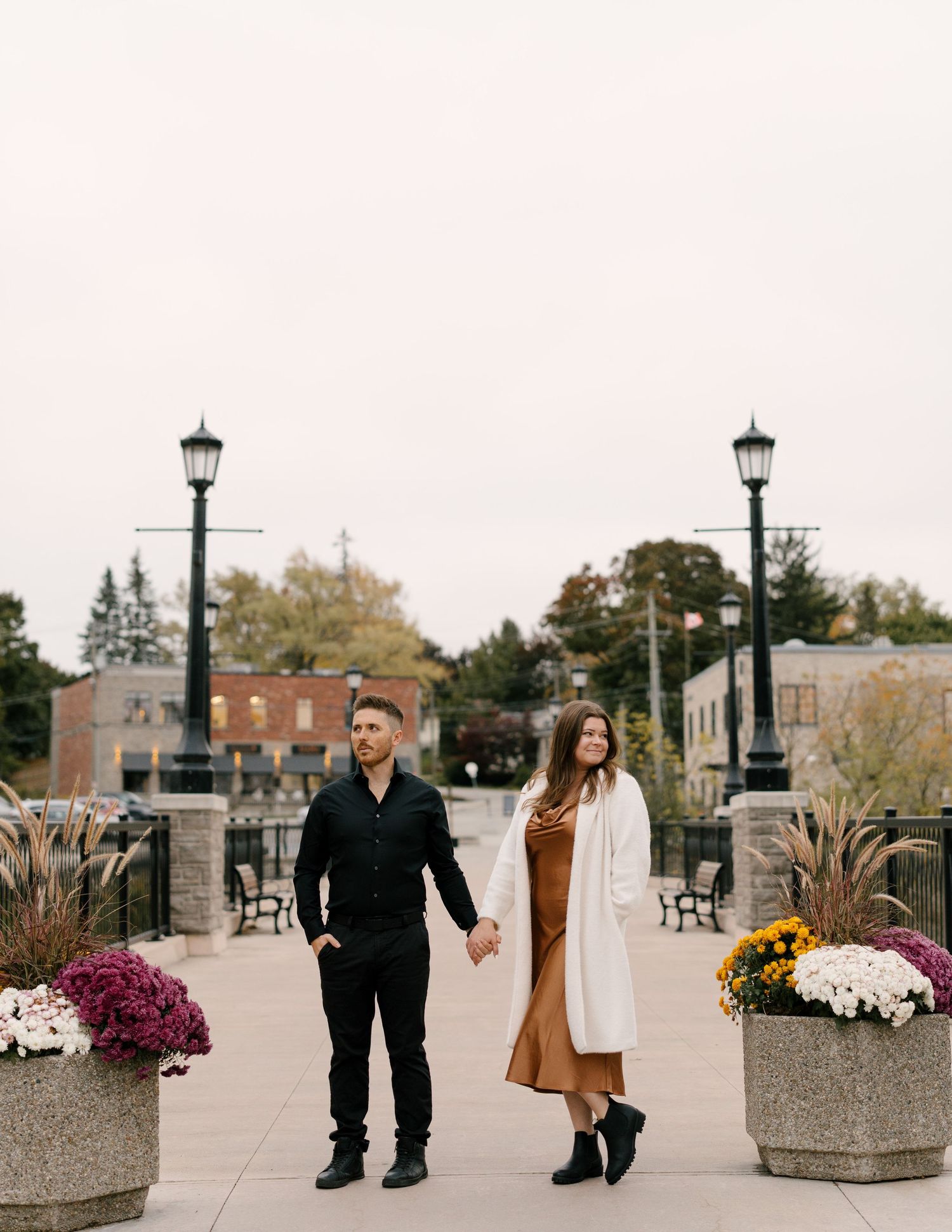 Elegant couple holding hands on a decorative bridge walkway surrounded by autumn flower planters.