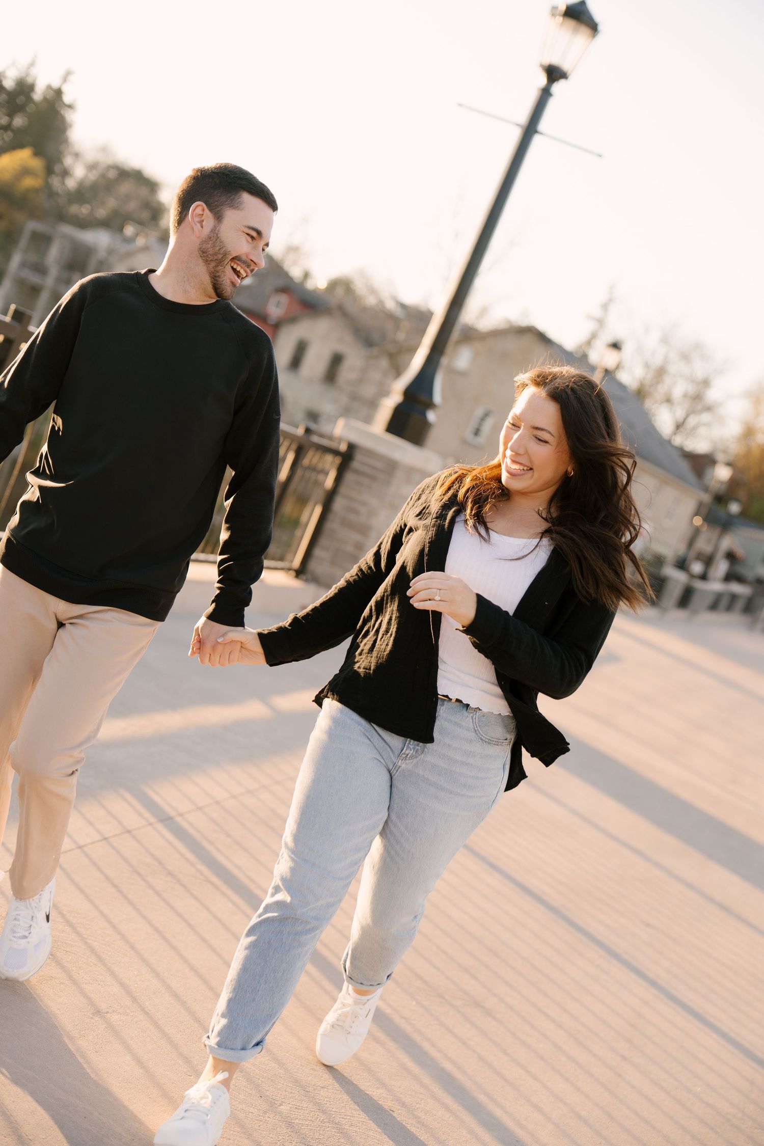 Happy couple laughing and holding hands while walking together on a sunny urban promenade in casual black outfits.