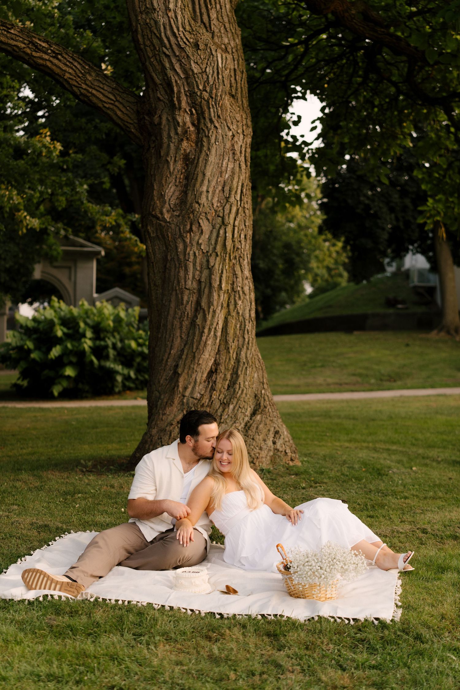 Couple relaxing on a white blanket under a large tree in a park during golden hour.