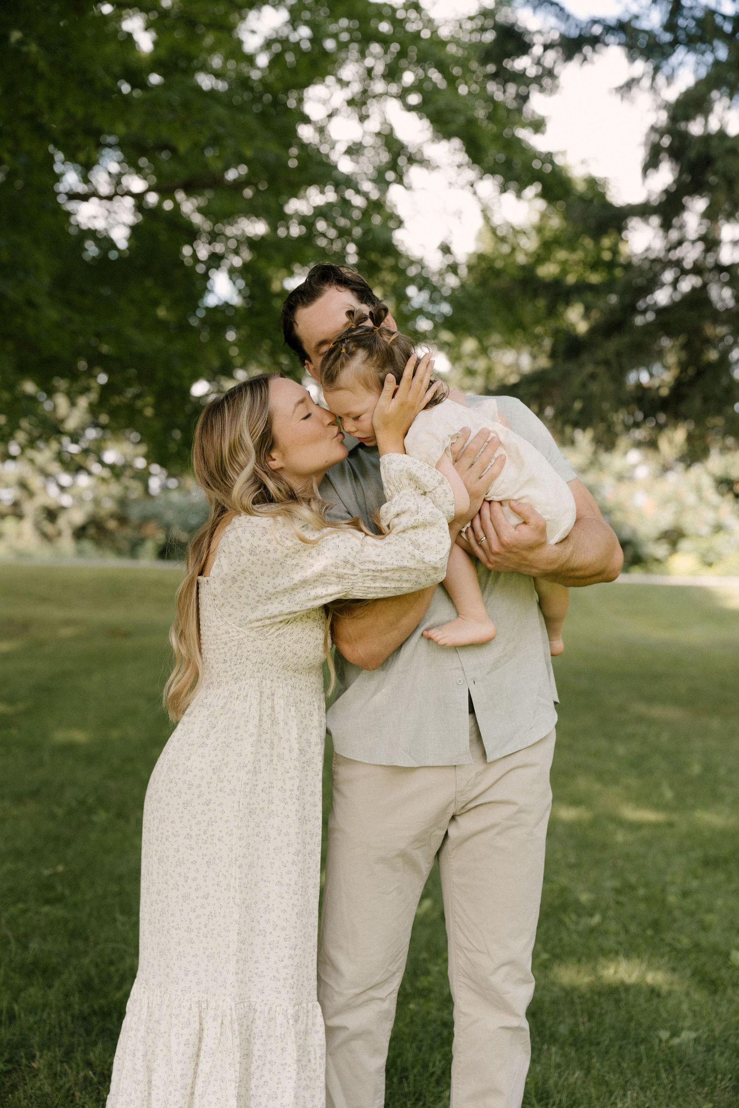 A couple in neutral tones kisses their baby girl in a lush green park setting.