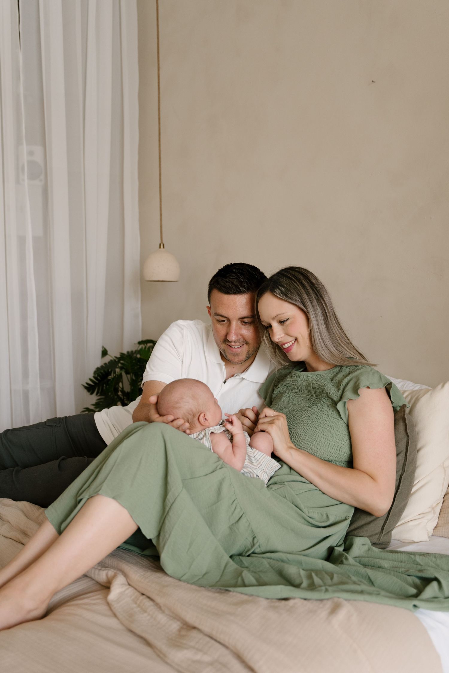 A happy couple sits on a bed smiling at their newborn baby, mother wearing a sage green dress.