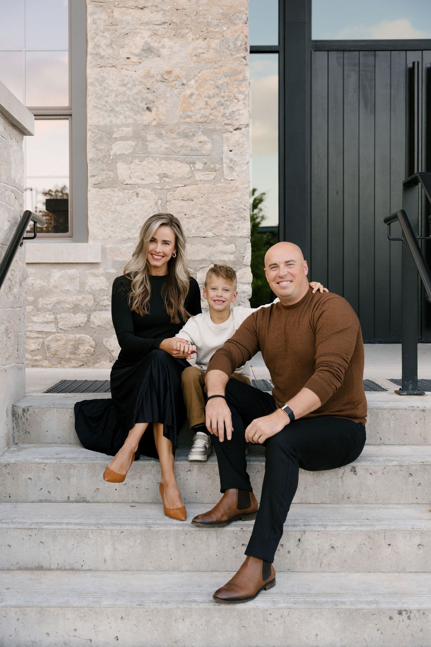 Happy family of three sitting on stone steps outside modern home entrance.