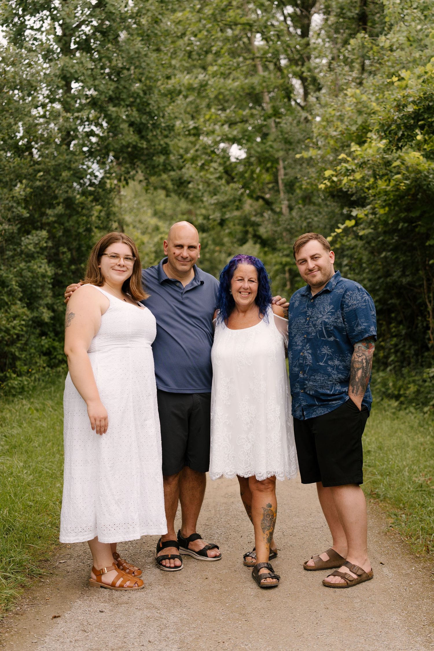 Family of four smiling together on a garden path, wearing casual summer outfits, surrounded by lush green trees.