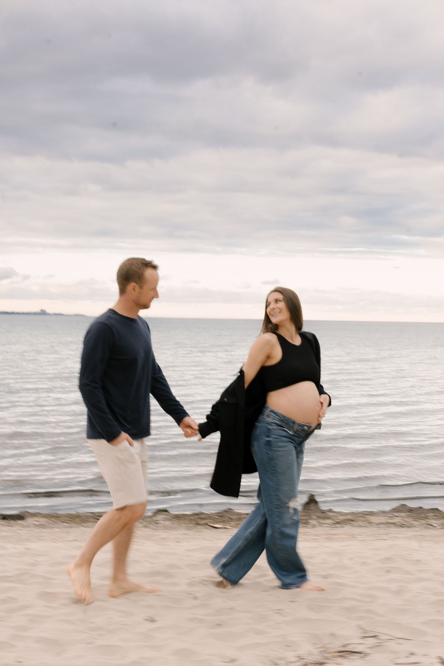 Pregnant woman in black outfit holds partners hand while walking barefoot on sandy beach at dusk.