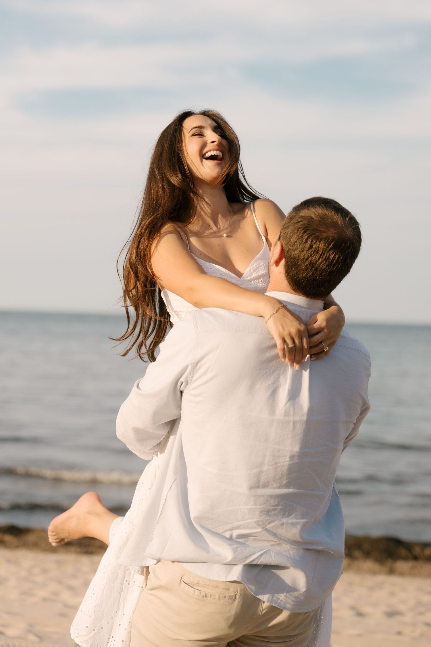 Man lifting joyful laughing woman in white dress on sandy beach with calm ocean in the background.