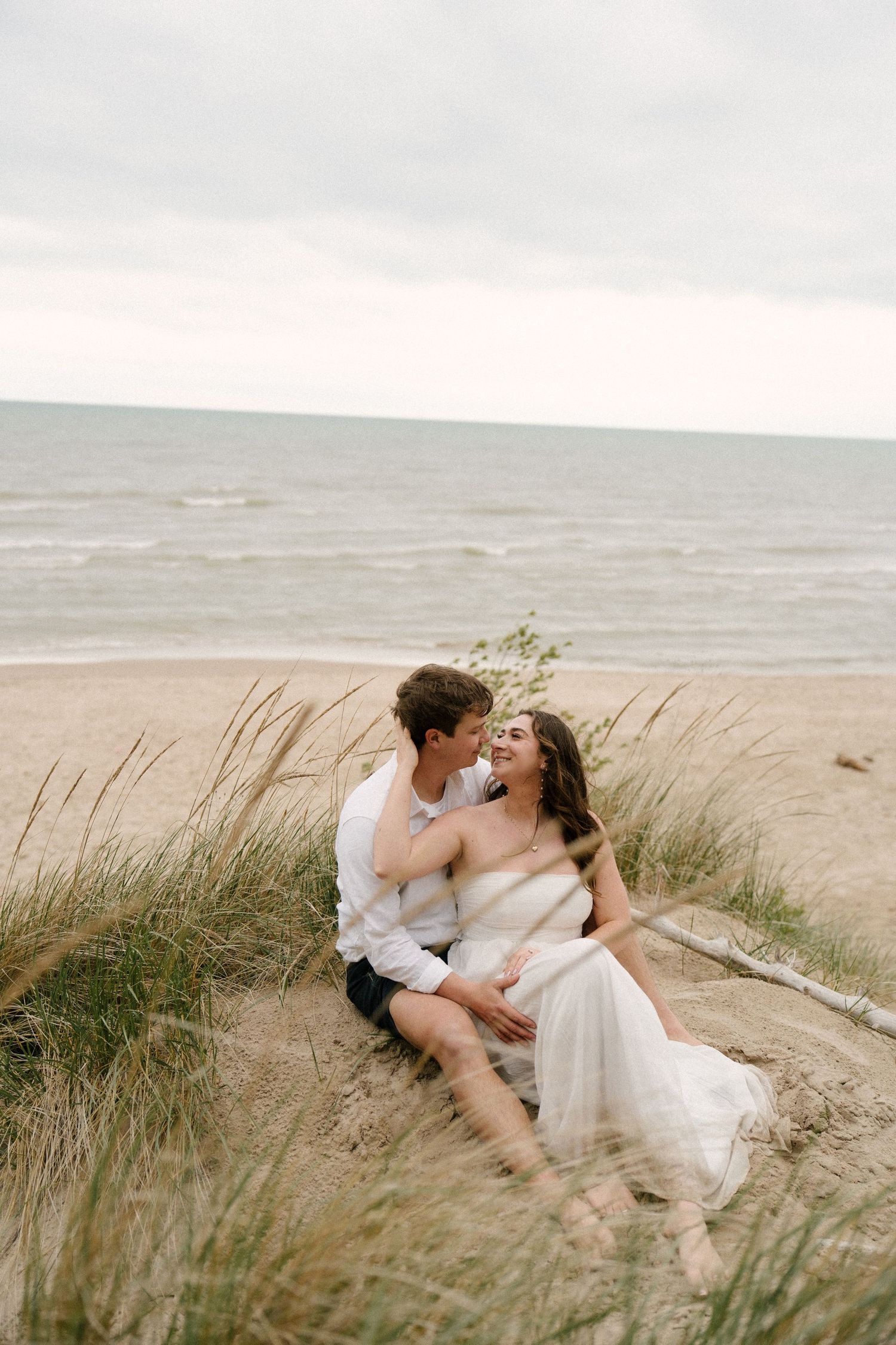 Couple in wedding attire sitting on sandy beach dunes, laughing and embracing with the ocean behind them.
