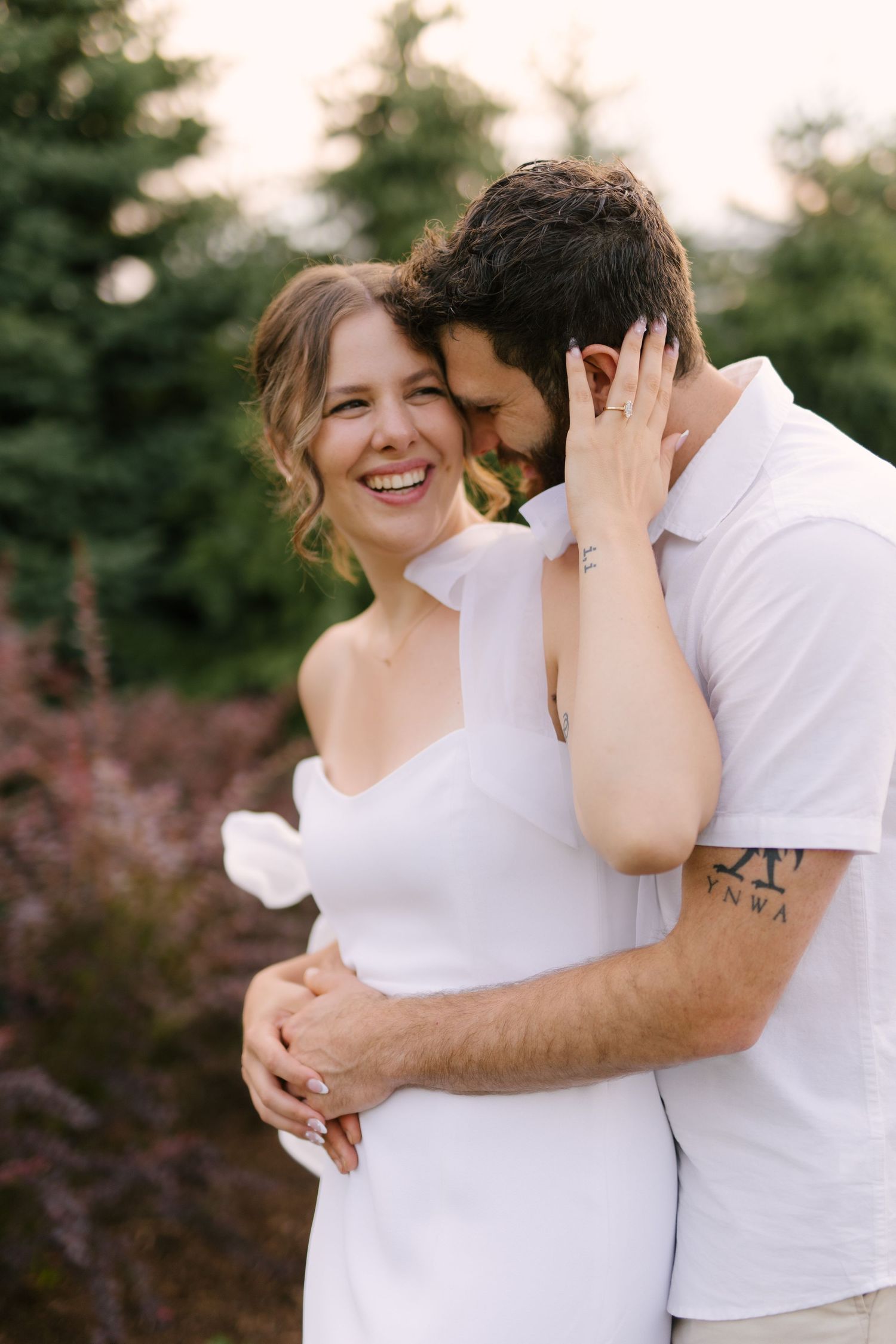 Happy couple cuddling in a garden, woman in white dress smiling while man with tattoo embraces her.
