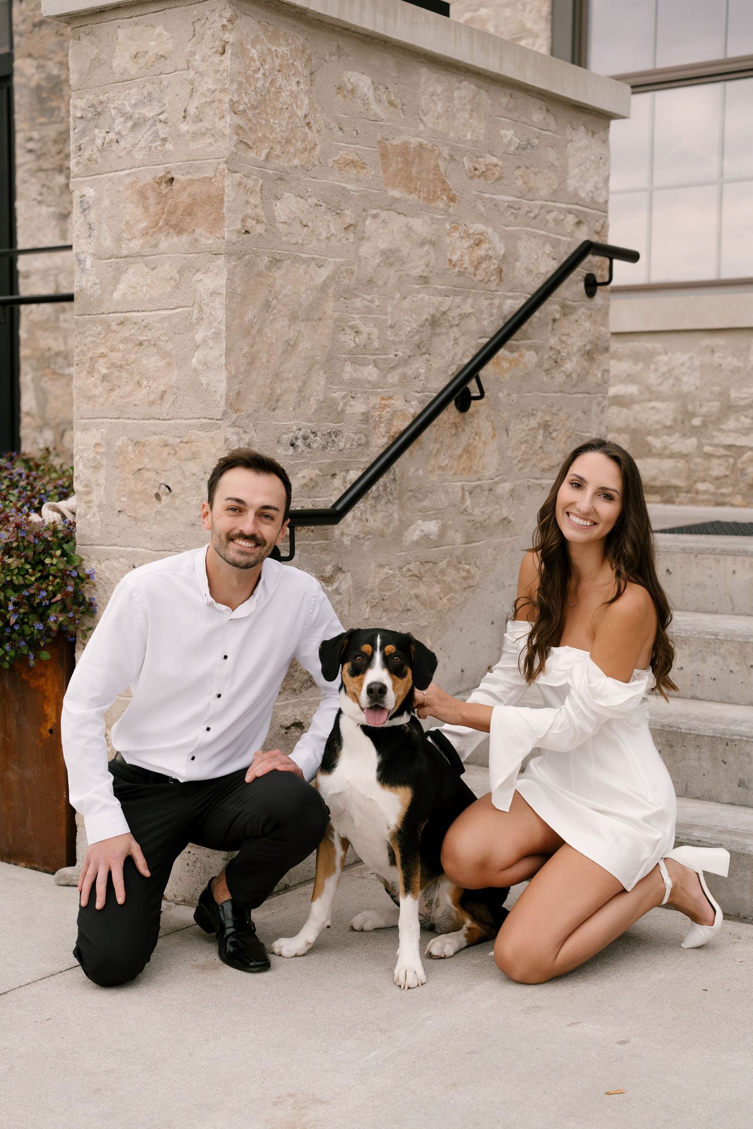 Smiling couple posing with their tricolor dog on steps outside a stone building.