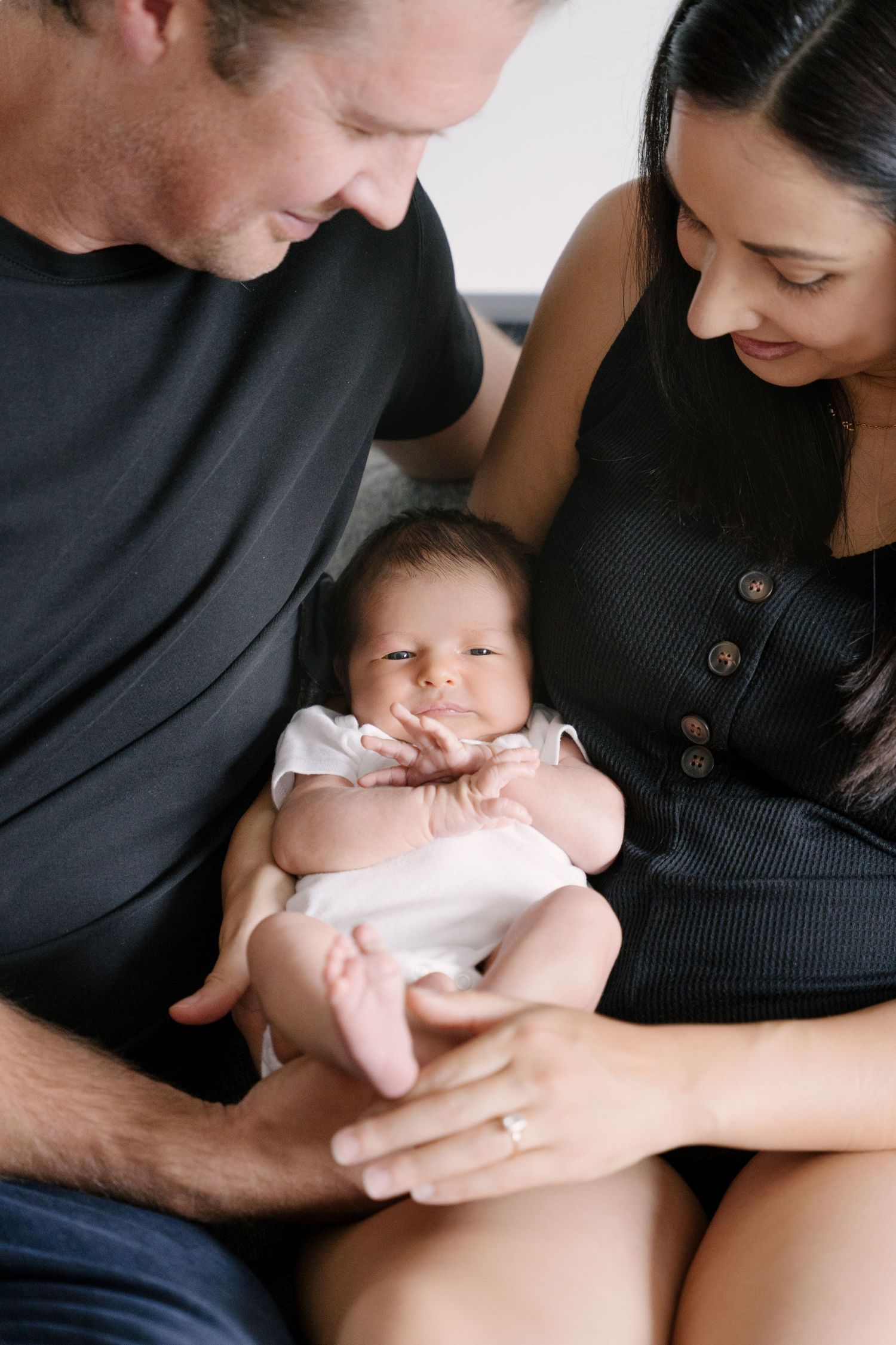 Parents in black outfits tenderly holding newborn baby together on a cozy couch.