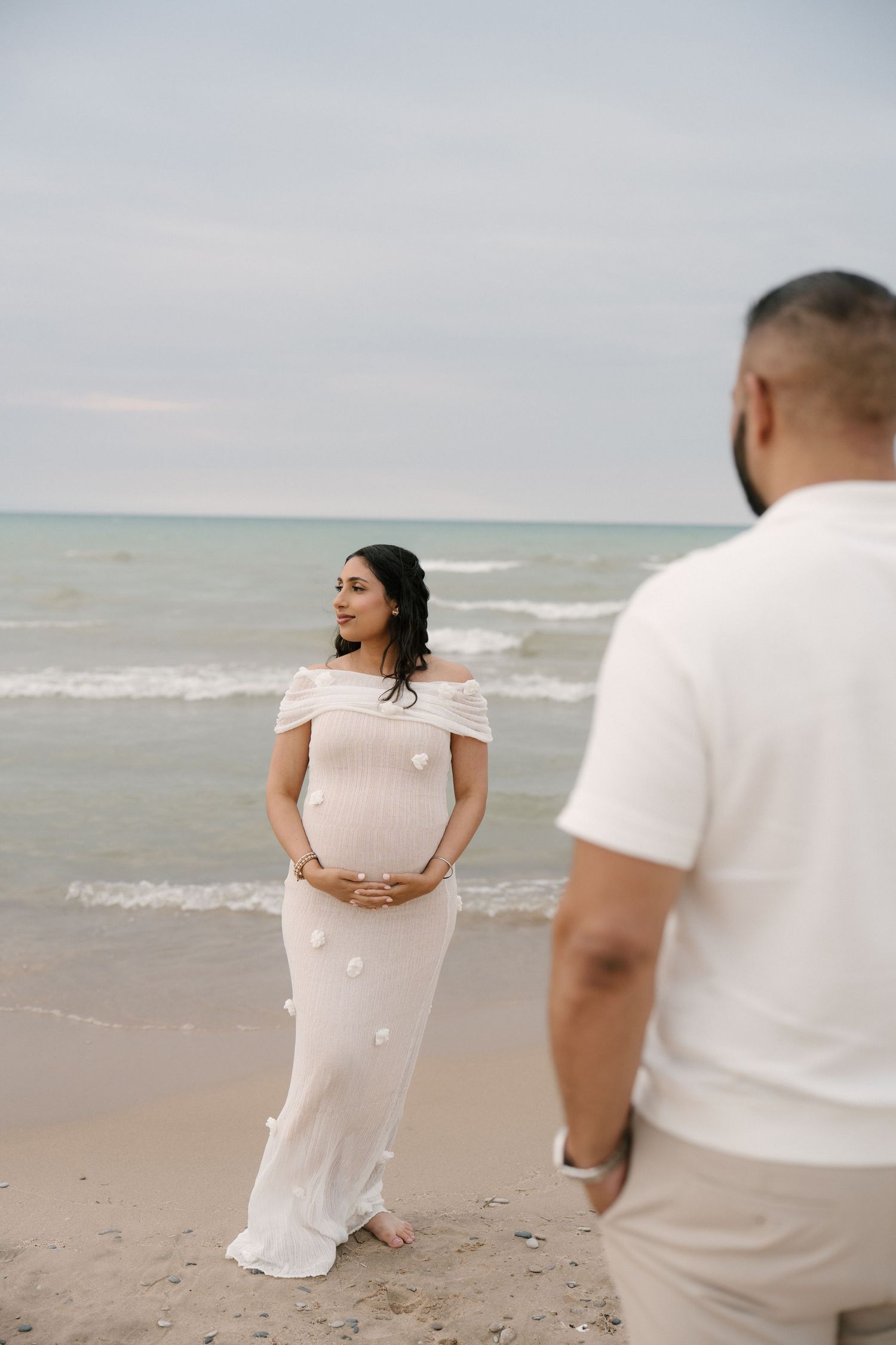 Pregnant woman in white off-shoulder dress smiles on beach as partner approaches from foreground.