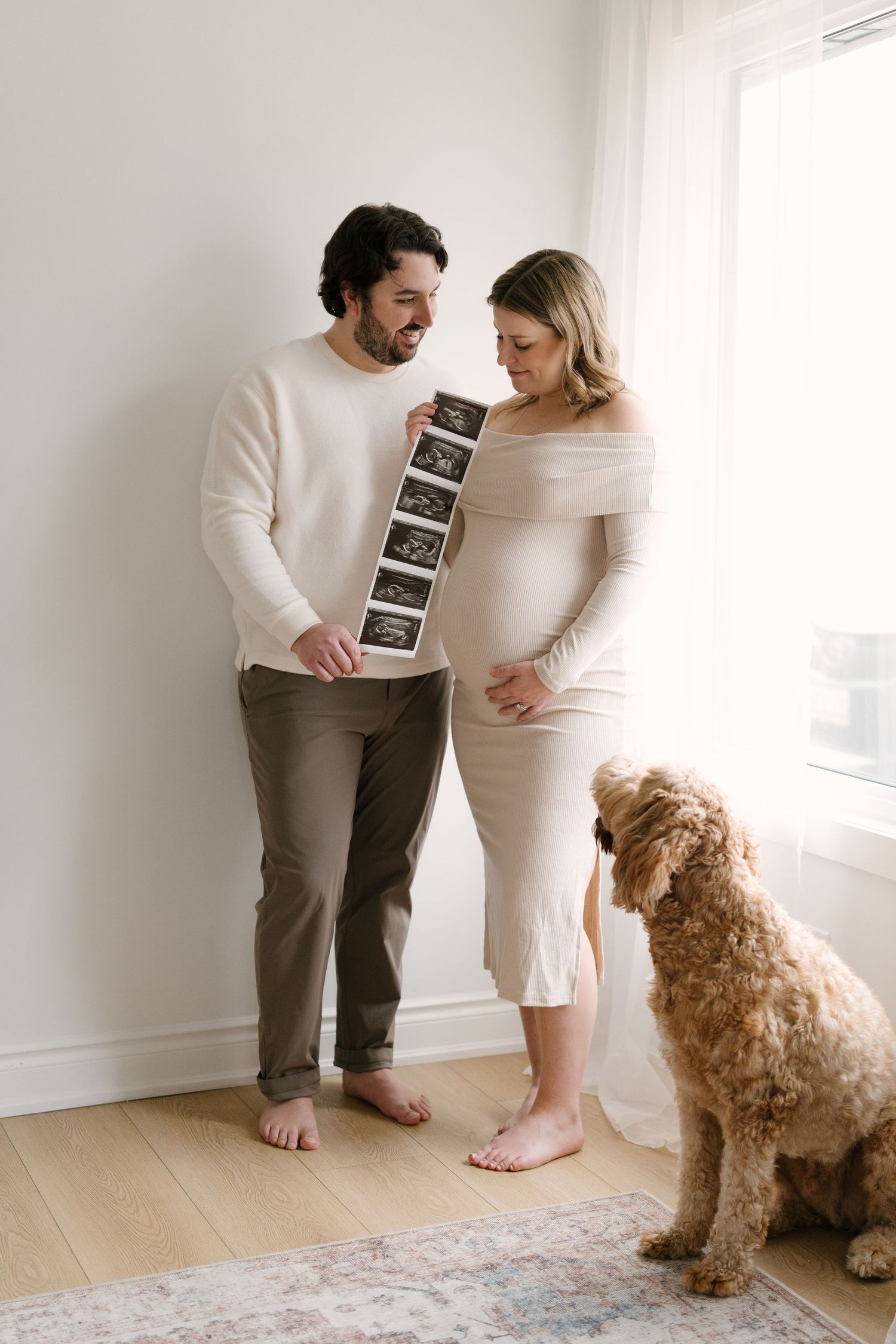 Expecting couple holding ultrasound photos with golden doodle dog beside them near a bright window.