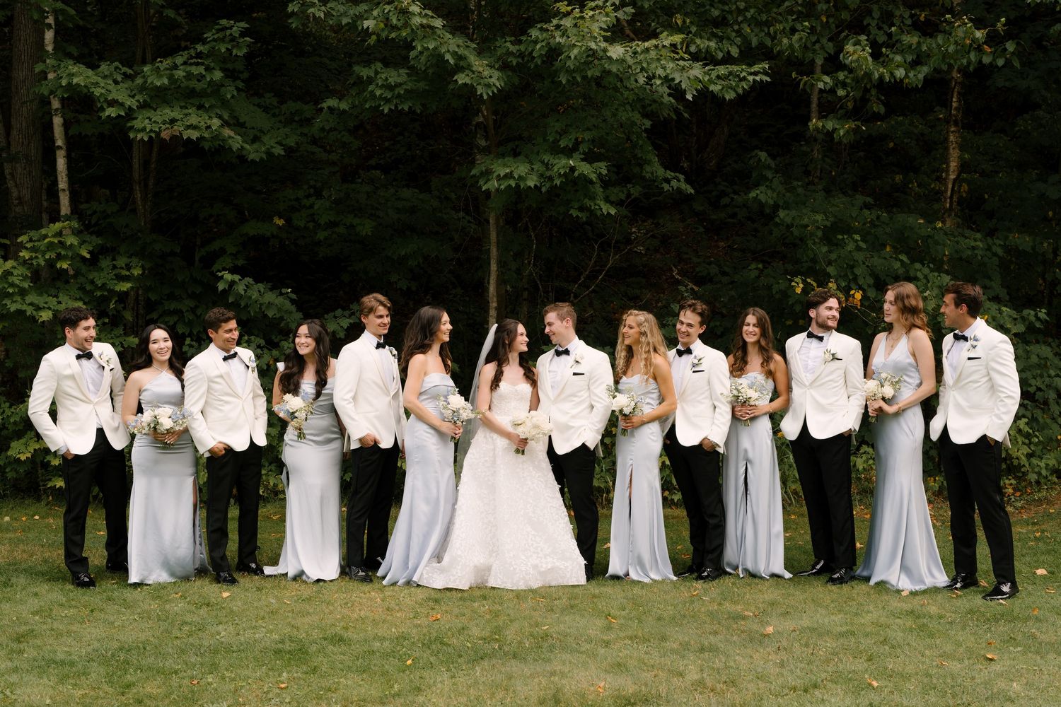 Wedding party poses outdoors, bridesmaids in blue gowns, groomsmen in black tuxedos, bride and groom centered before lush forest.