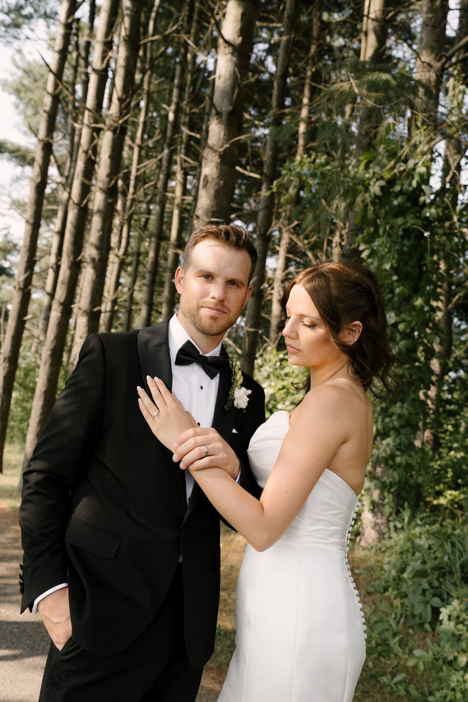 Groom in black tuxedo stands with bride in strapless white gown holding his lapel among tall pine trees.