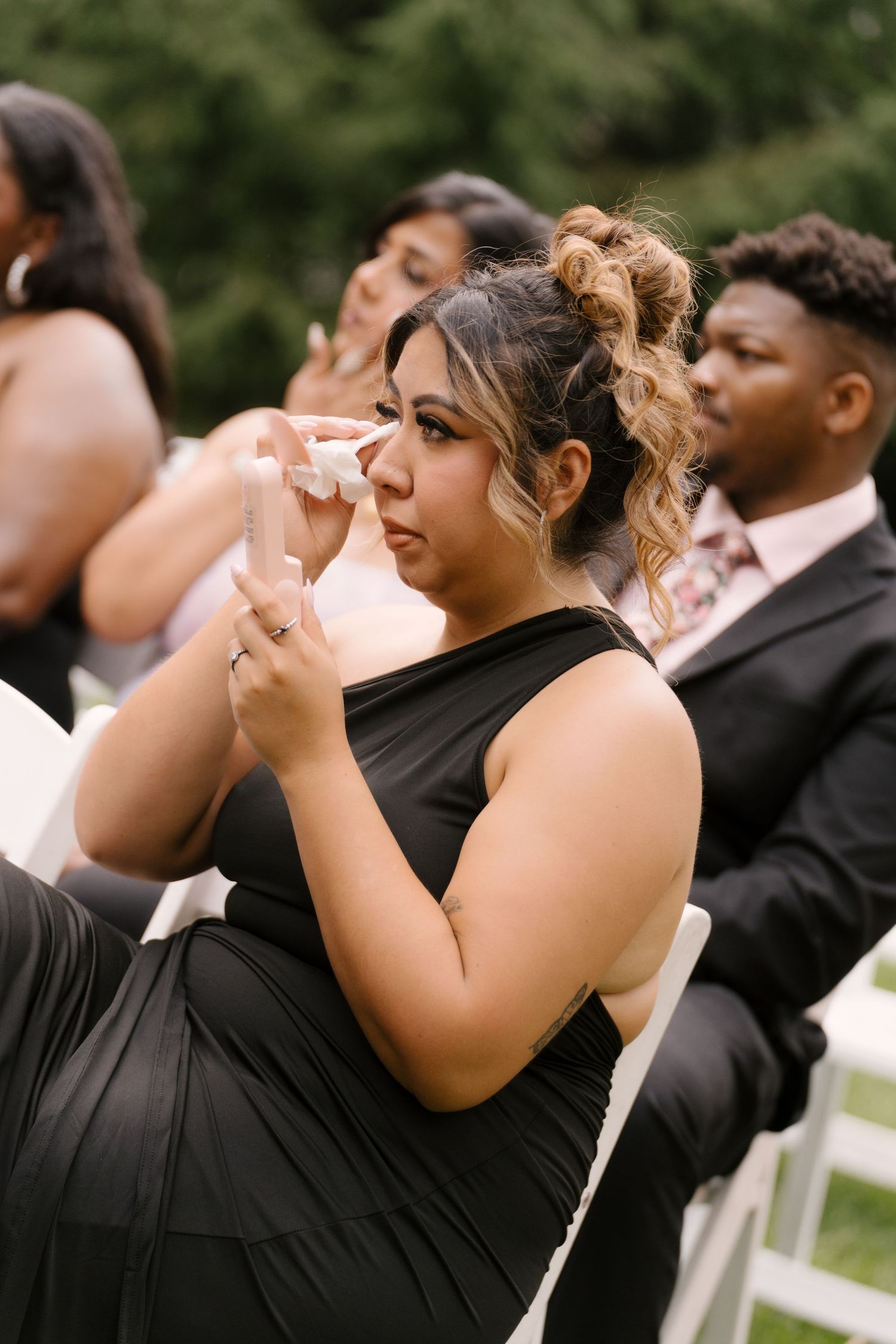 Emotional wedding guest in black dress dabbing tears with tissue during outdoor ceremony.