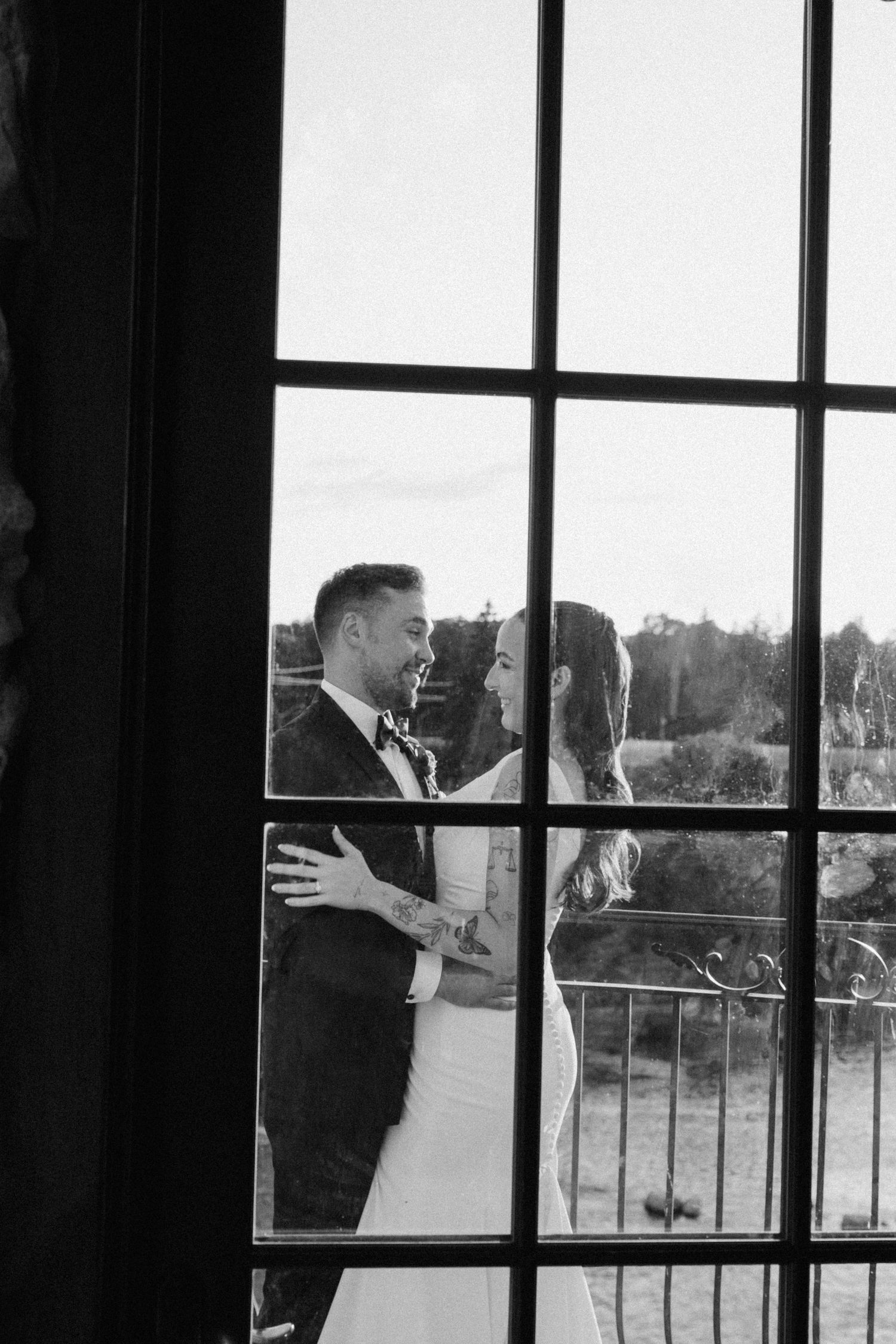 Bride and groom share intimate moment on balcony viewed through large window panes in black and white at Cambridge Mill wedding veune.