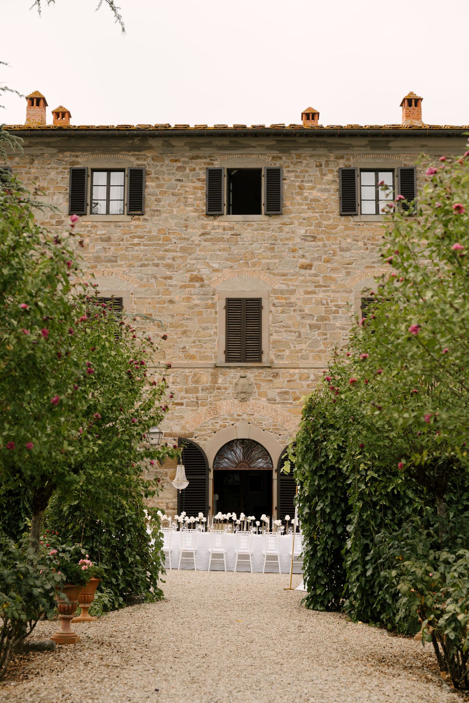 Stone Tuscan villa wedding venue with gravel path lined by lush green trees and white chairs.