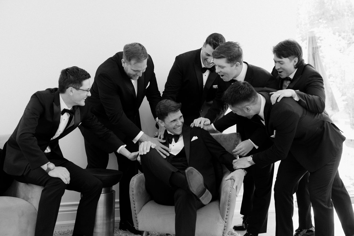 Groomsmen in black tuxedos huddling around groom seated in chair in candid black and white wedding photo.