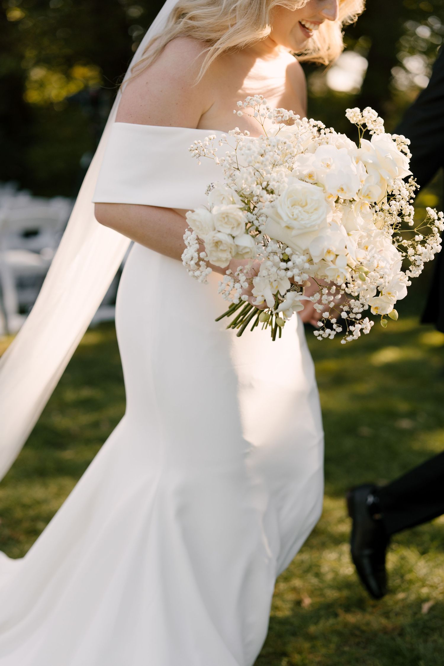 Bride in off-shoulder mermaid gown carries elegant white rose and baby's breath bouquet outdoors.