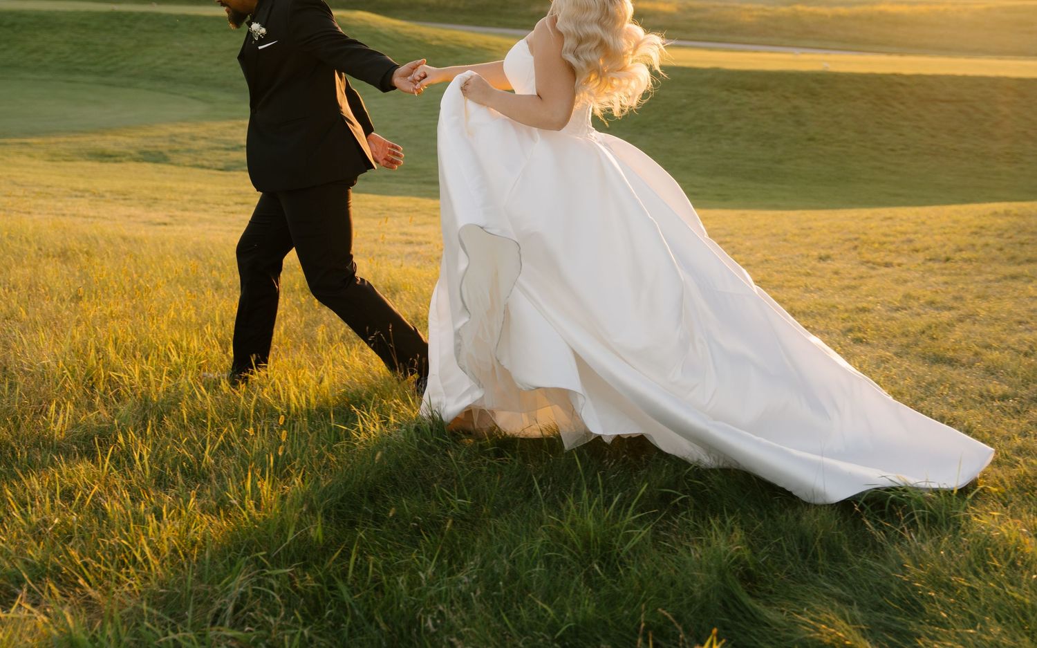 Bride in flowing white wedding gown running through lush green grass with groom at golden hour.