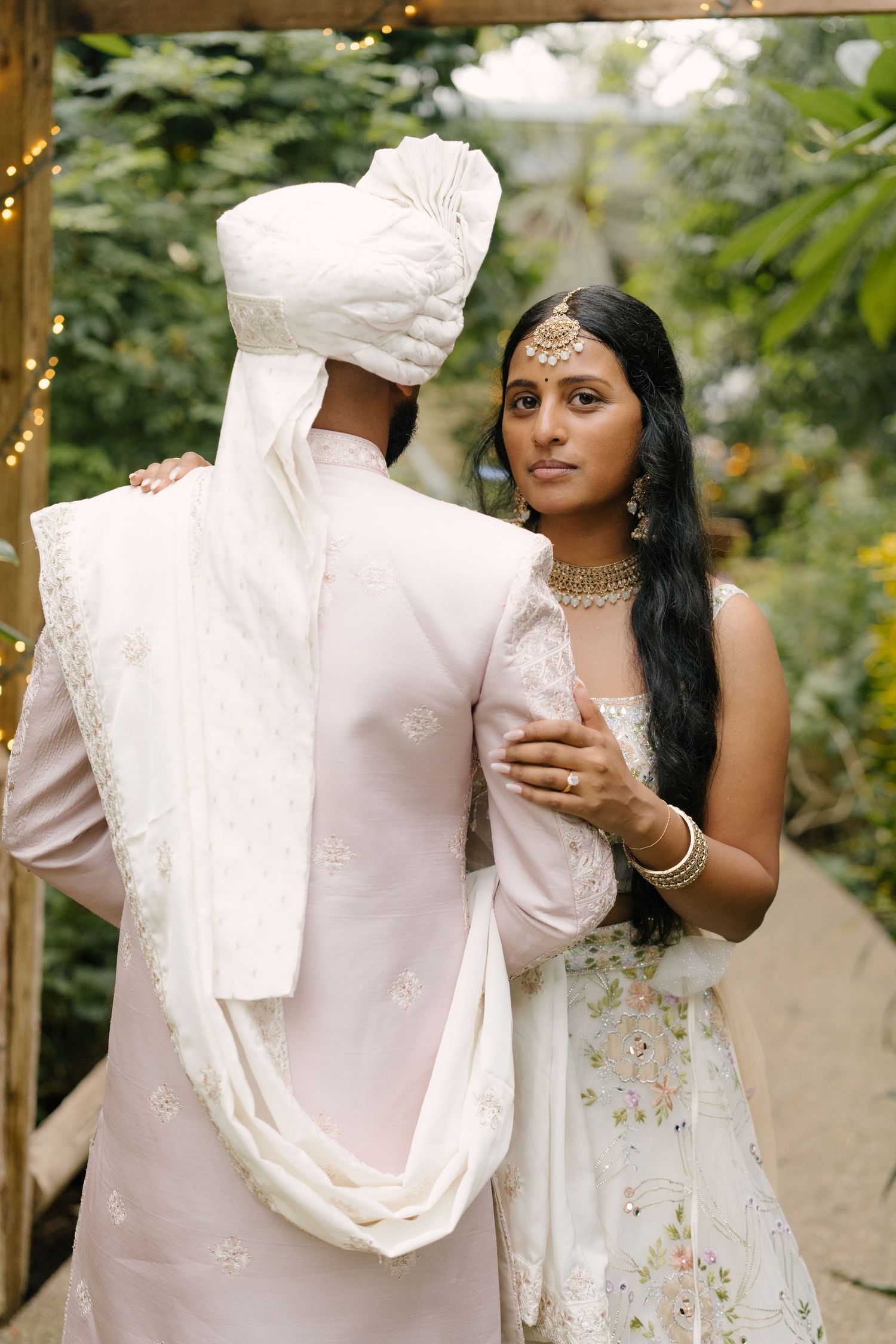 Indian bride in white lehenga gazes at camera while groom in turban faces away.