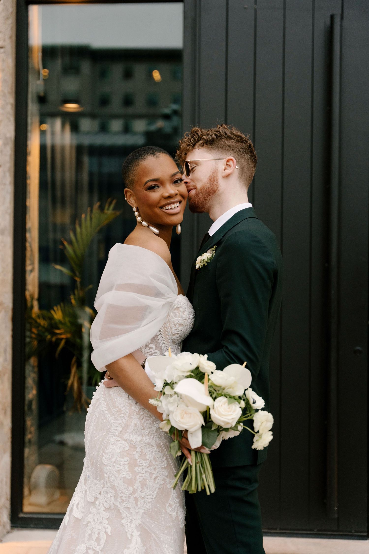 A joyful interracial couple poses on their wedding day, bride in lace gown holding white bouquet as groom kisses her cheek.