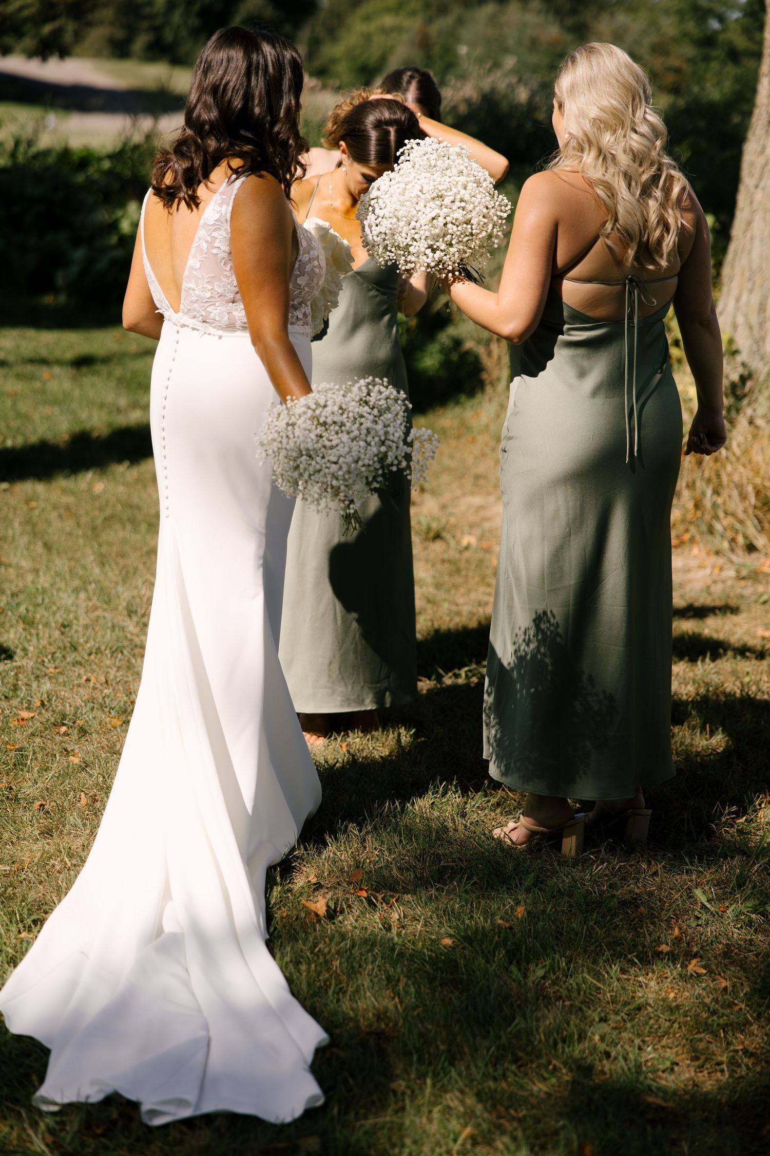 Bride and bridesmaids in sage dresses stand together outdoors holding white baby's breath bouquets.