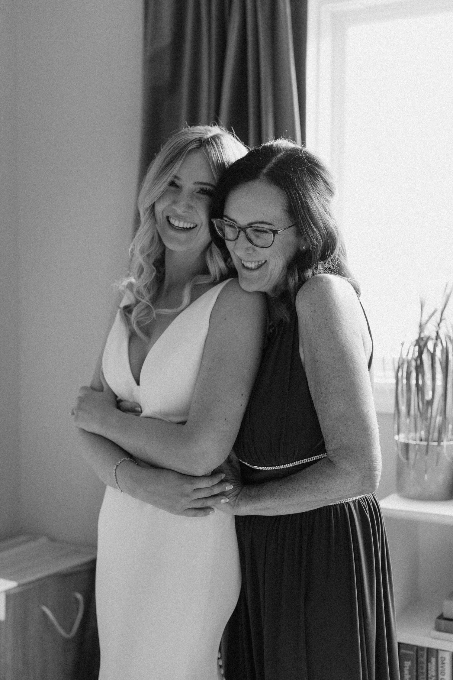 Black and white photo of a bride in a white gown sharing a joyful hug with a woman in a dark dress.