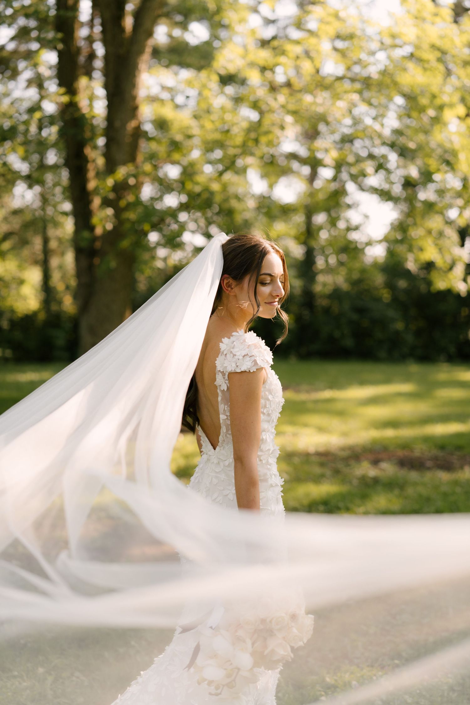 Smiling bride in a floral lace gown poses outdoors as her long white veil flows in the wind.