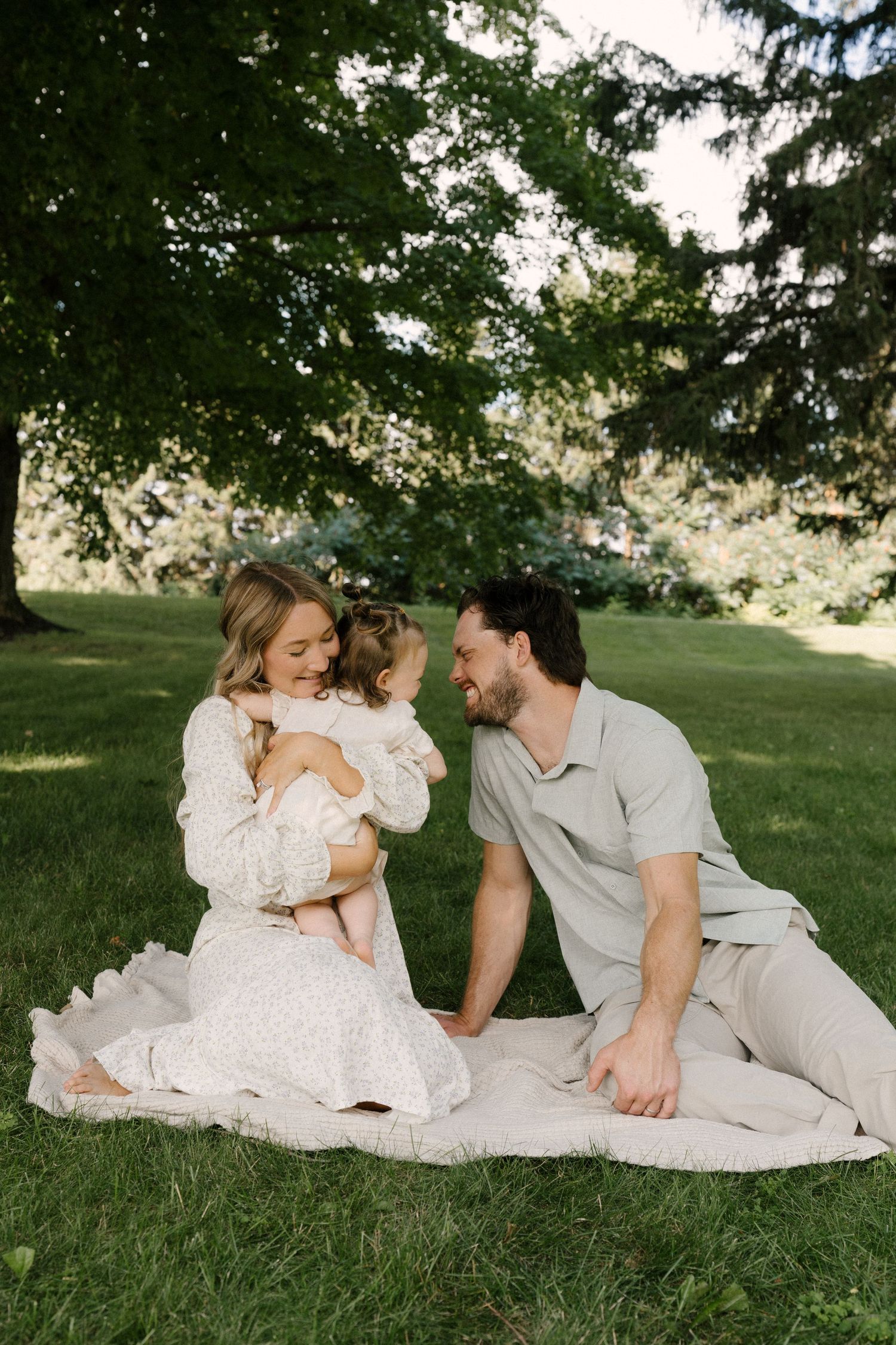 Happy young family sitting on blanket under large tree in lush green park with baby.